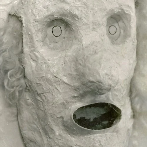 A close-up of a sculpture of a face with exaggerated features and large, circular eyes, made from a rough, textured material covered in white paint or plaster.