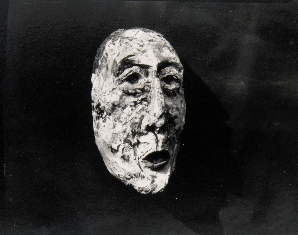A black and white photo of a face mask made from layered paper, resembling an elderly person's face, floating against a dark background.