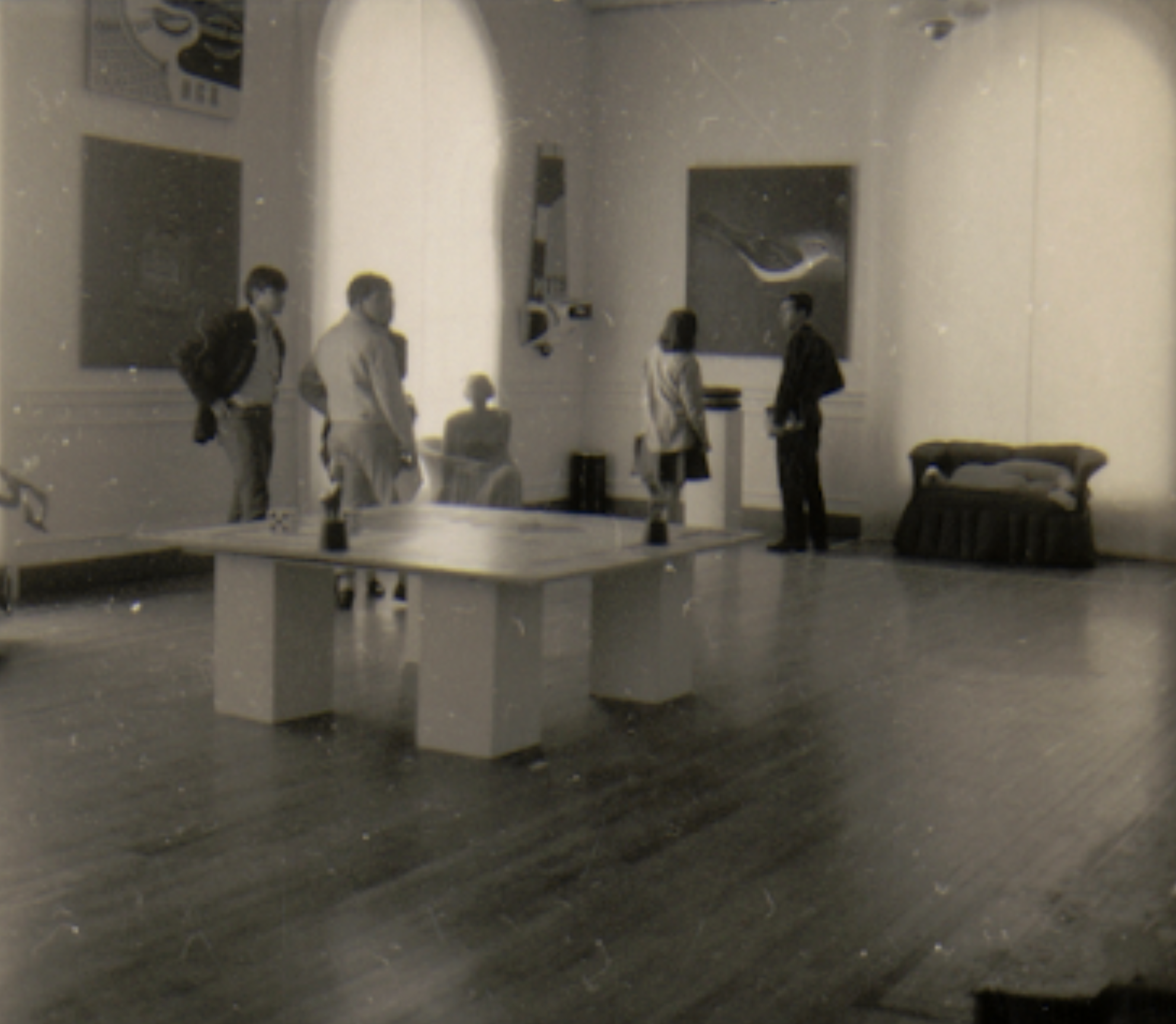A black-and-white photo of a cafe with five people, some standing and some sitting, and a table with drinks in the foreground.