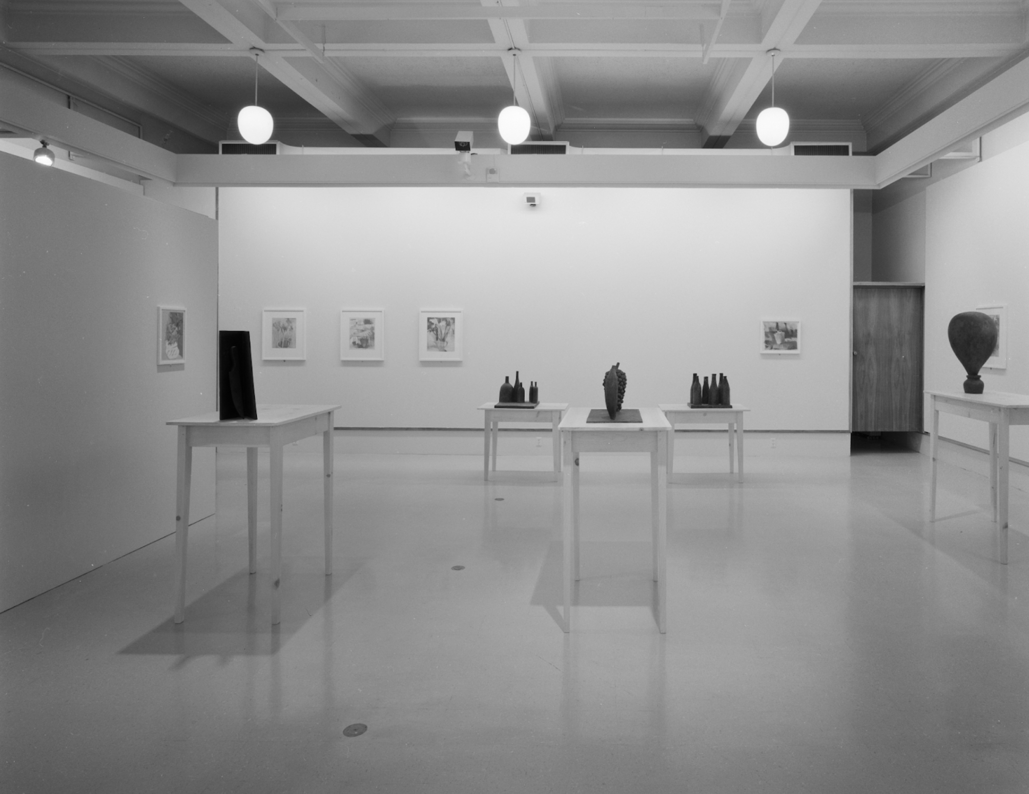 Black and white photo of an art gallery with framed artwork on walls and sculptures on individual tables.