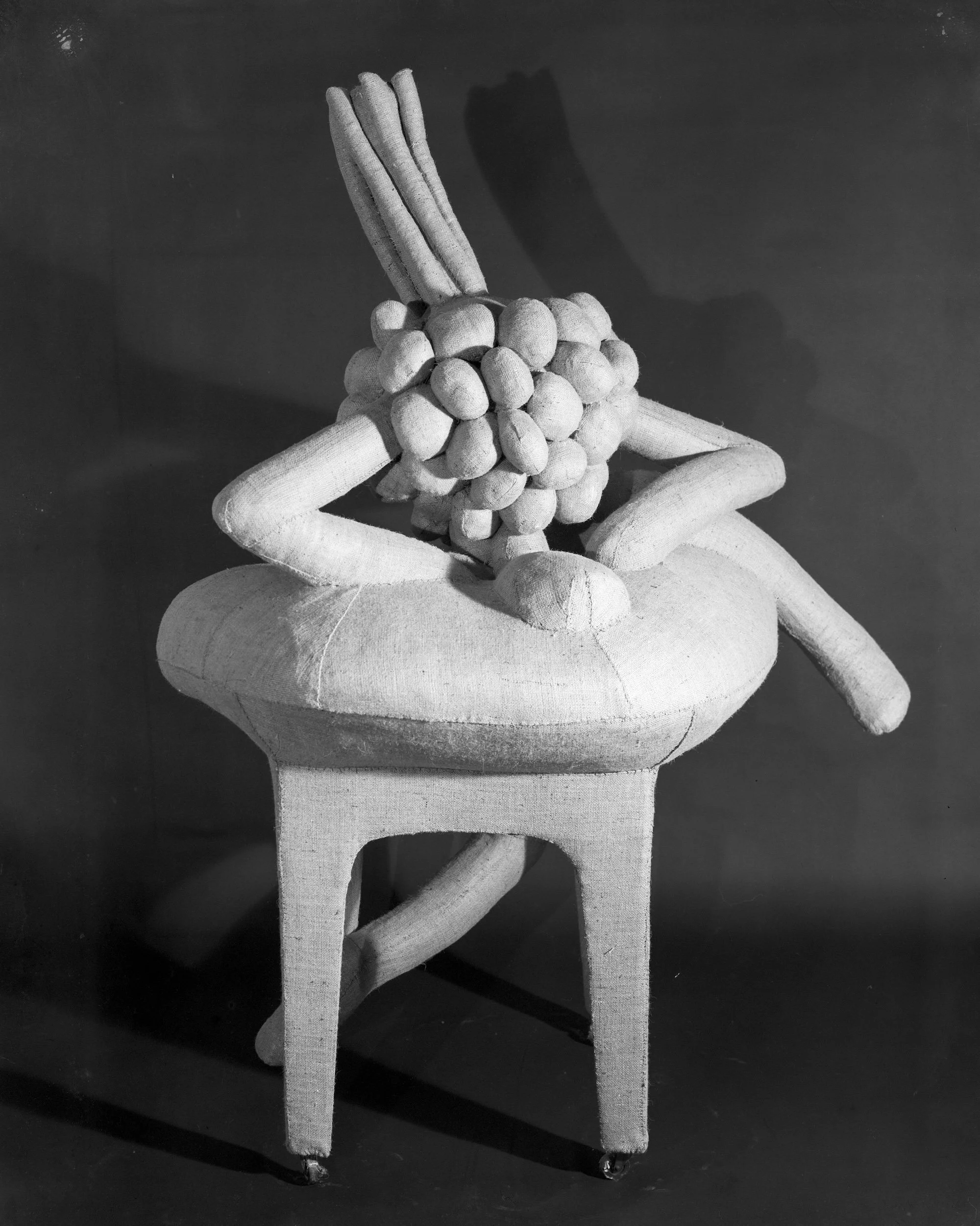 A wooden chair with a rounded seat and four legs, with an art sculpture on it made of various chunks of wood assembled to resemble a fruit or cluster, with elongated wooden pieces extending upward.