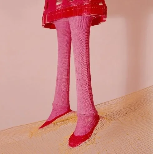 An art sculpture of a pair of pink legs with pink fishnet stockings and red shoes, beneath a red cage-like structure, set on a yellow mesh surface.