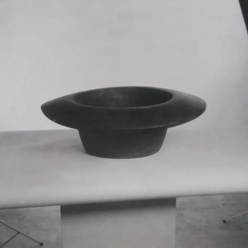 A black ceramic vessel with a wide, flat rim and a narrow base, placed on a white surface against a white background.