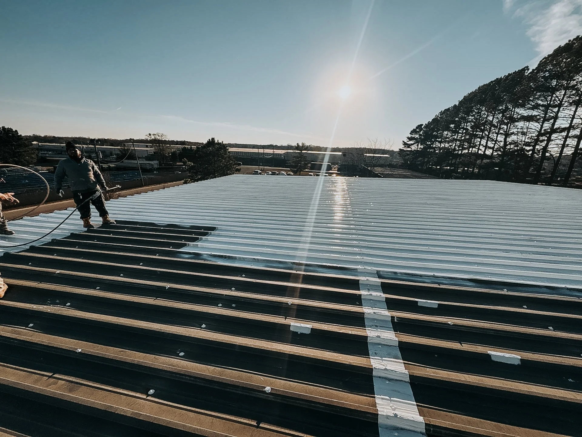 Workers installing metal roofing on a building during sunny weather with trees and power lines in the background.
