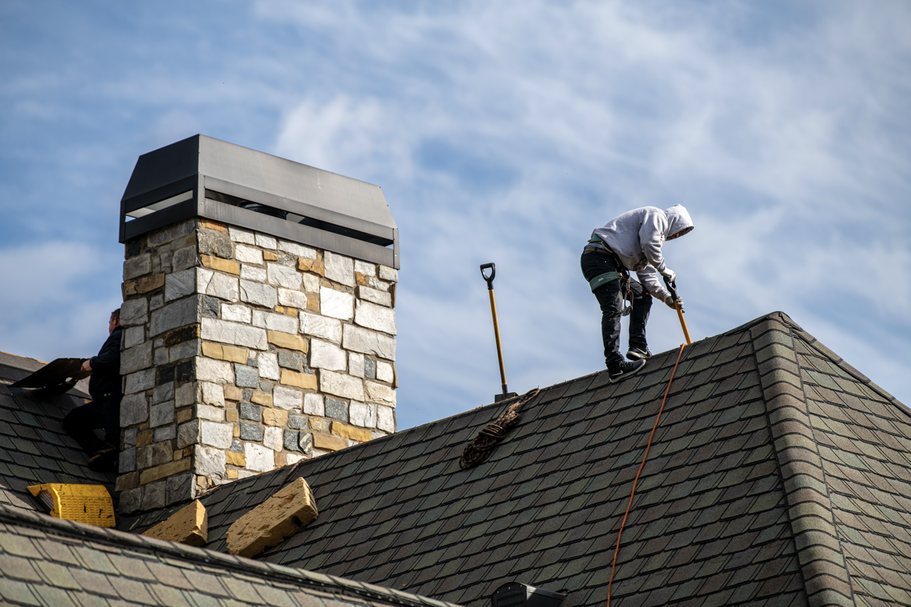 Worker cleaning or repairing a roof while standing on shingles, with a chimney nearby under a blue sky.