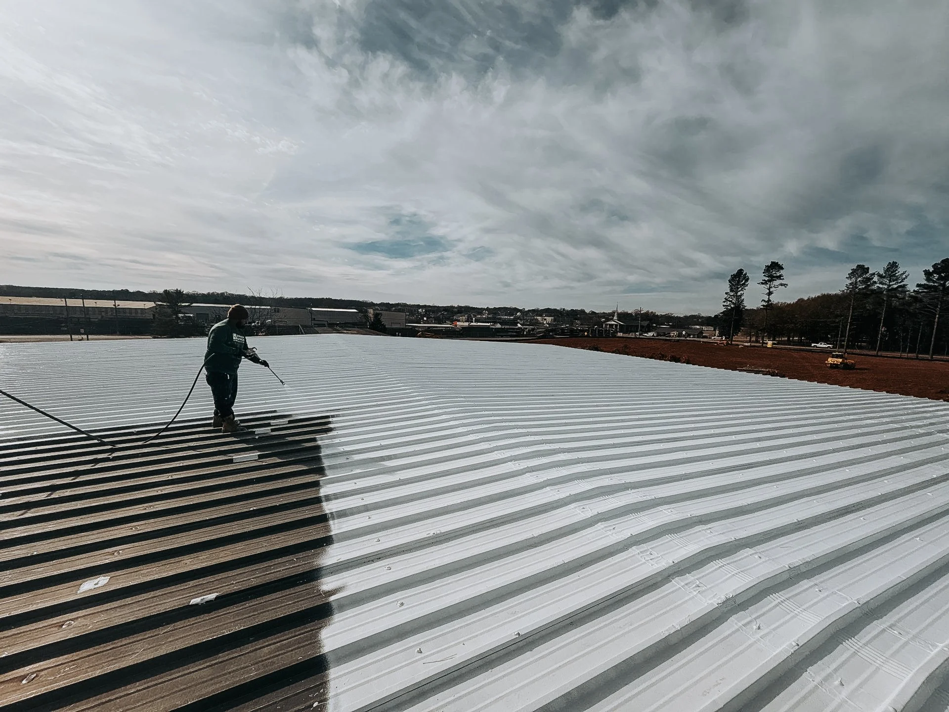 A worker sprays white paint on a large corrugated metal roof, with part of the roof still unpainted. The sky is cloudy with some blue showing, and trees and buildings are visible in the background.