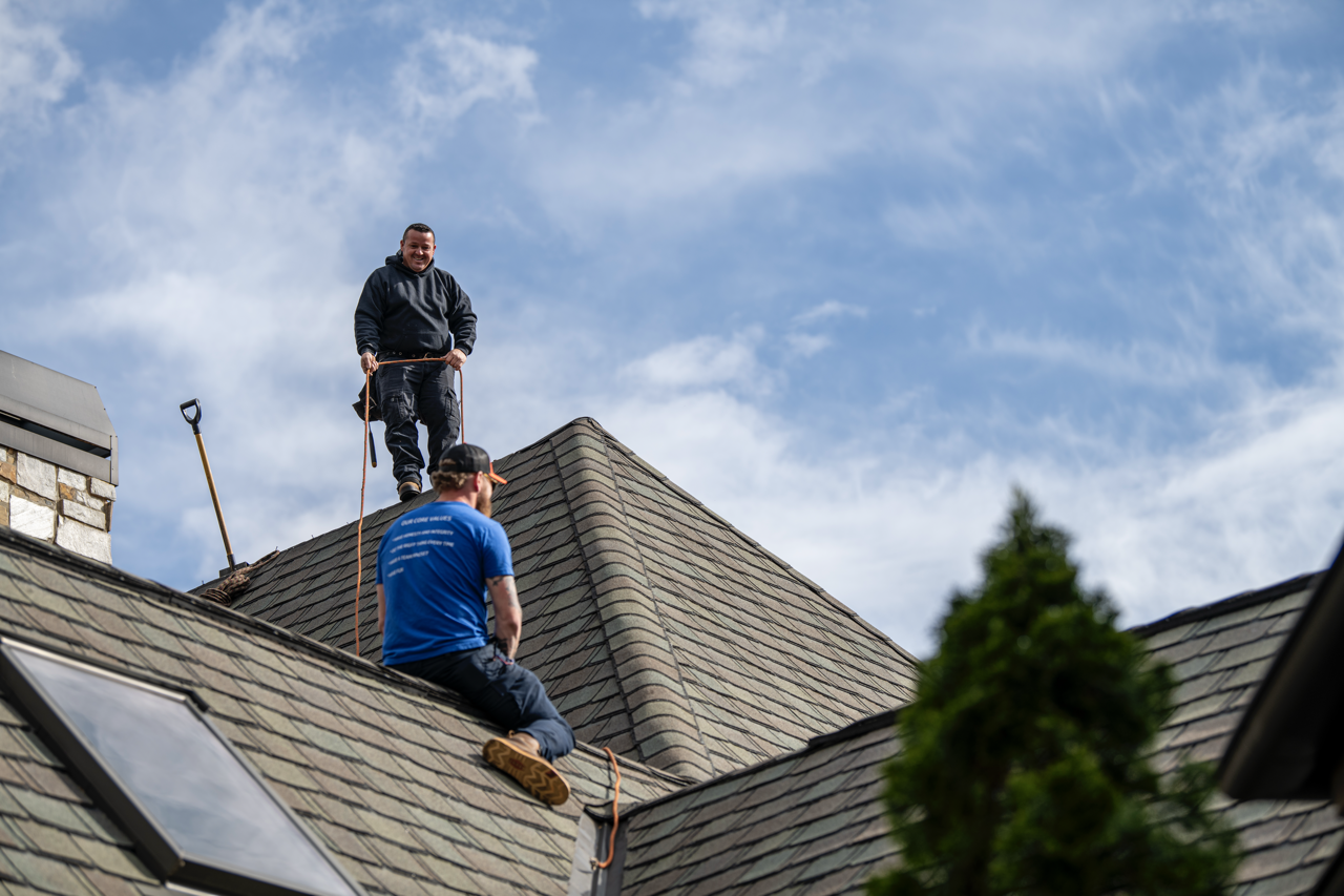 Two workers on a house roof, one standing and holding a safety harness and the other sitting, with a blue sky and clouds in the background.