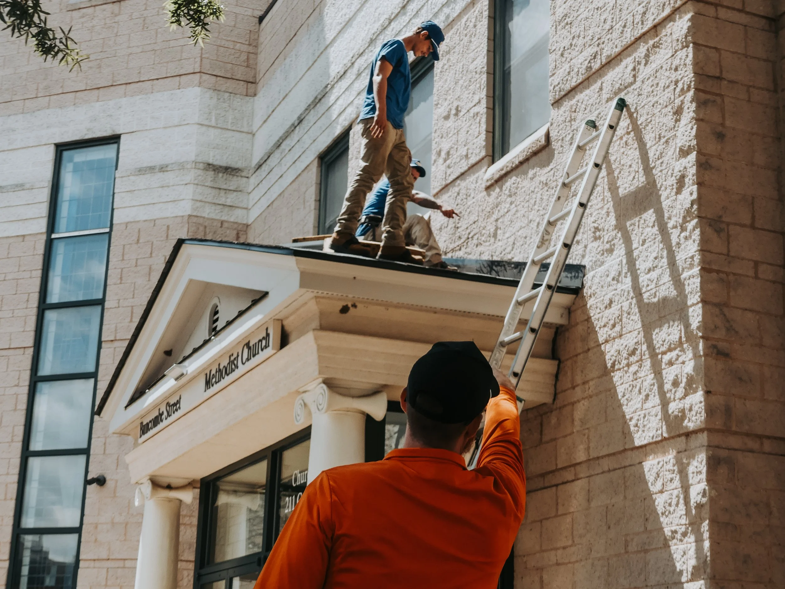 Three men working on the roof of a building with two of them standing and one sitting. One man is climbing a ladder. The building has a sign indicating it is a Methodist Church.