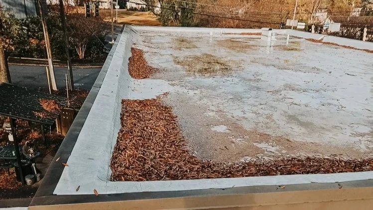 A flat rooftop covered with leaves, dirt, and debris in some areas, with a roof edge visible in the foreground and trees and power lines in the background.