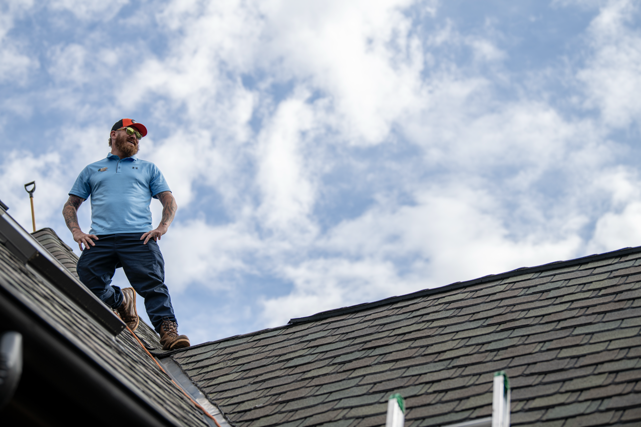 Man standing on a roof wearing sunglasses, a cap, a blue polo shirt, and work boots, with a cloudy sky in the background.