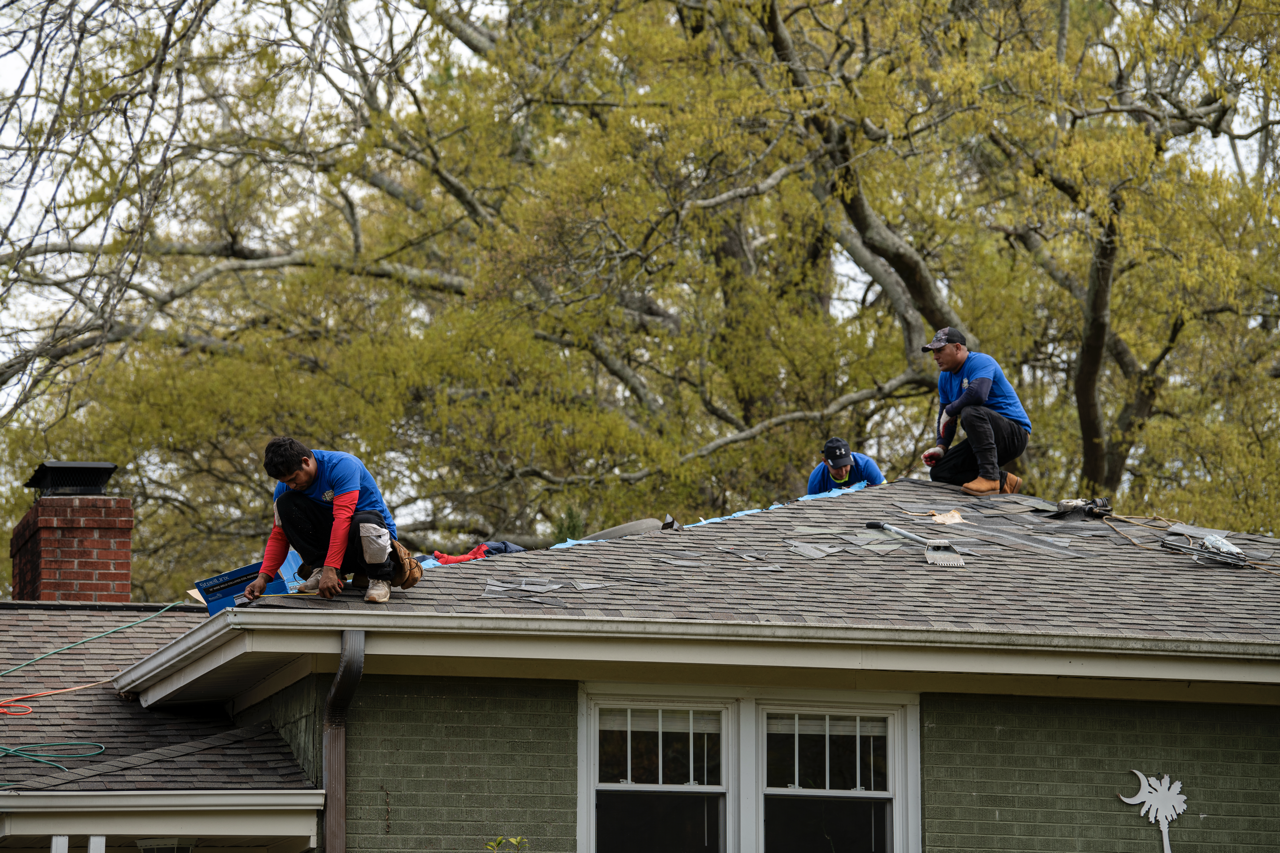 Three workers installing new shingles on a house roof while sitting on the shingles, with trees in the background.