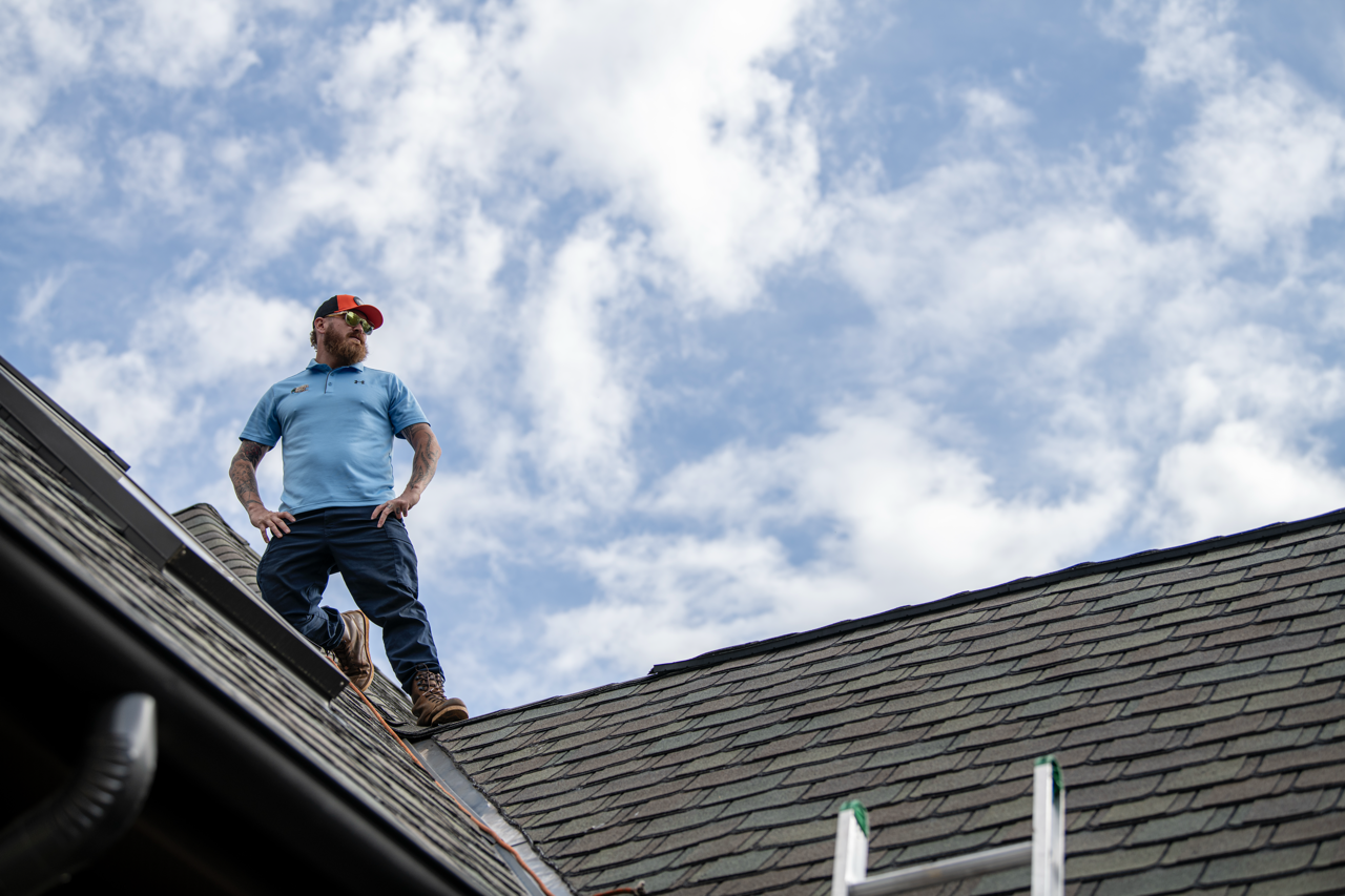 A man wearing a blue shirt, dark pants, and a red cap stands on a roof, looking into the distance against a cloudy sky.