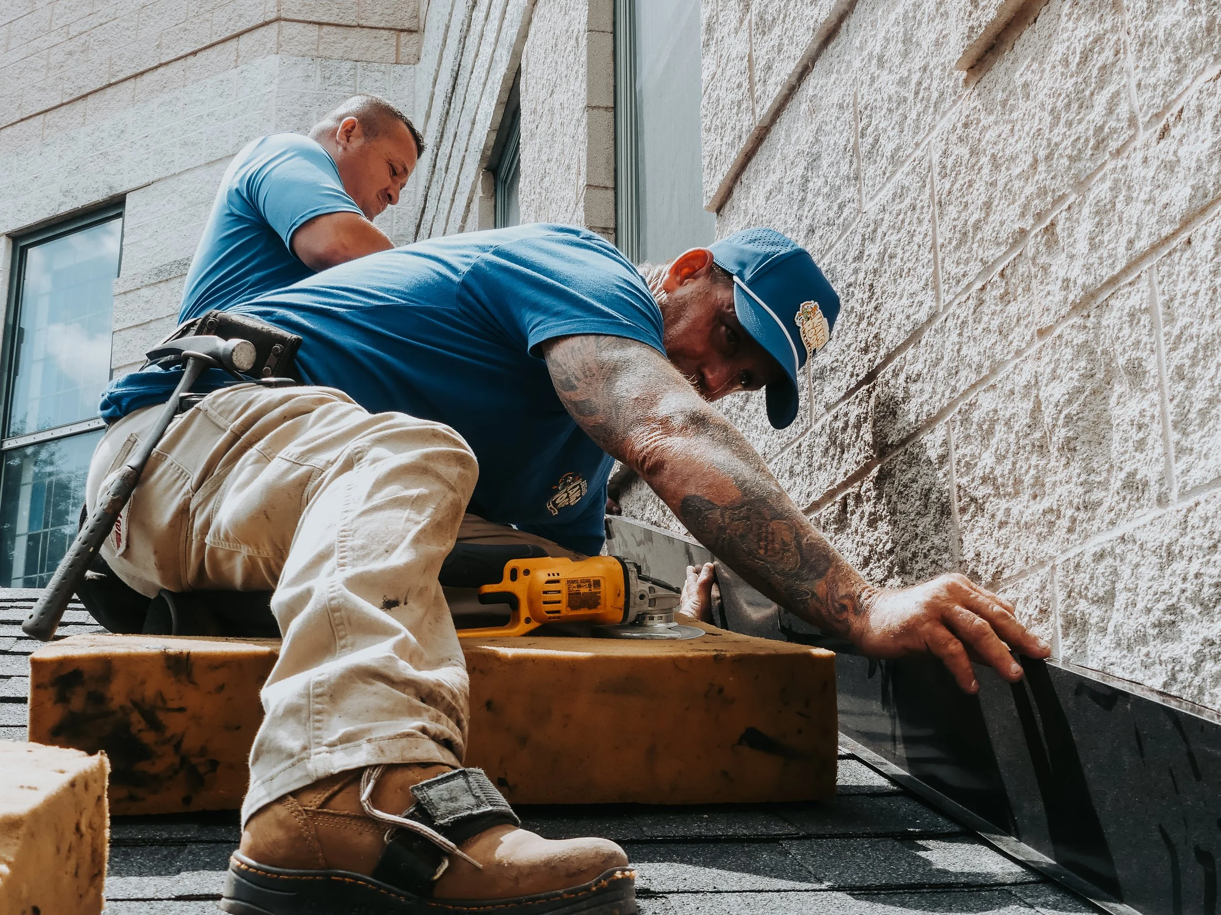 Two construction workers installing flashing on a building's roof. One worker is kneeling and using a power tool, while the other is standing and observing.