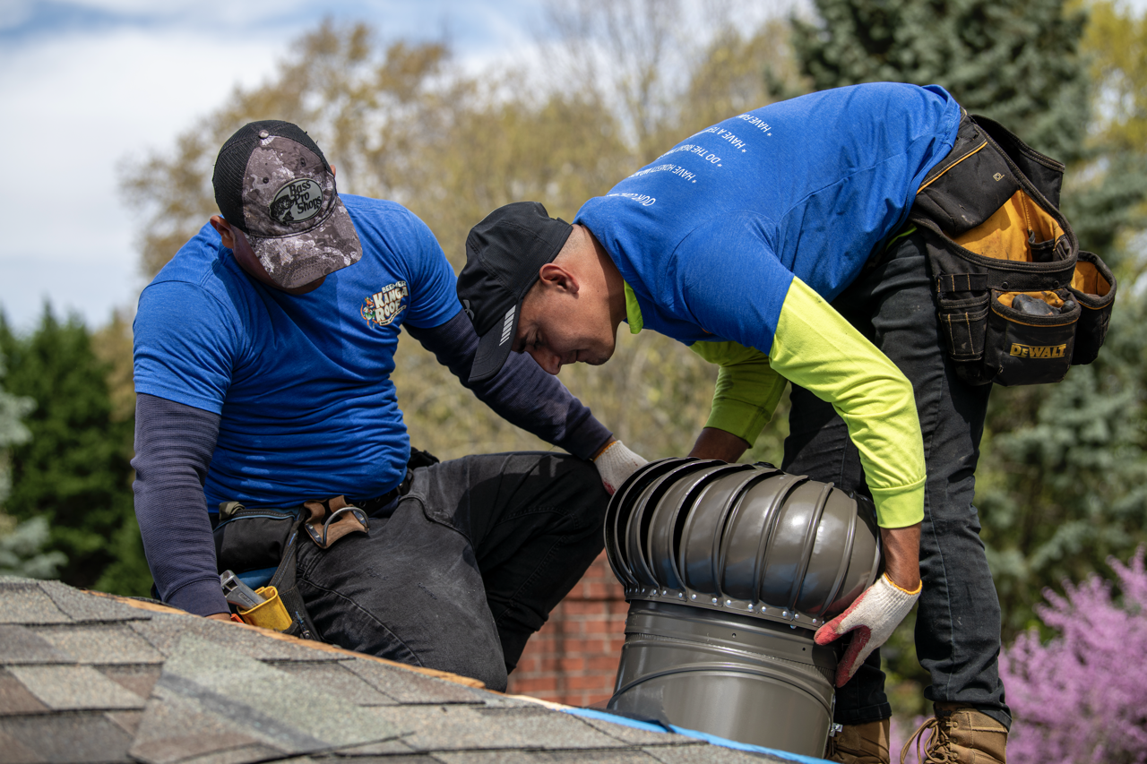 Two workers installing a new metal vent on a roof, surrounded by trees and greenery.