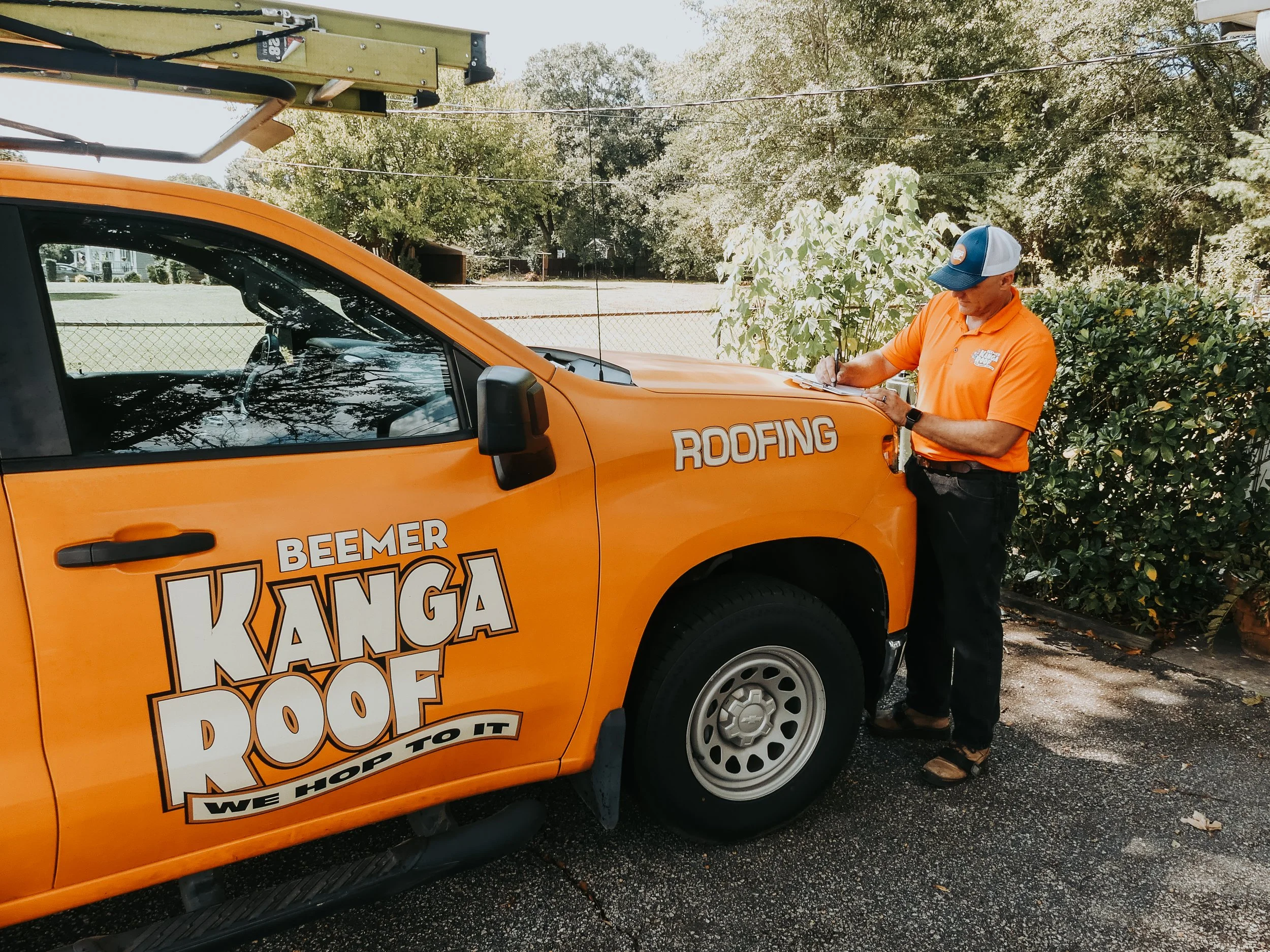 A worker in an orange shirt, wearing a cap, is standing next to an orange roofing company truck, writing on a clipboard. The truck has company branding, including the words 'Beemer Kanga Roof' and 'Roofing'.