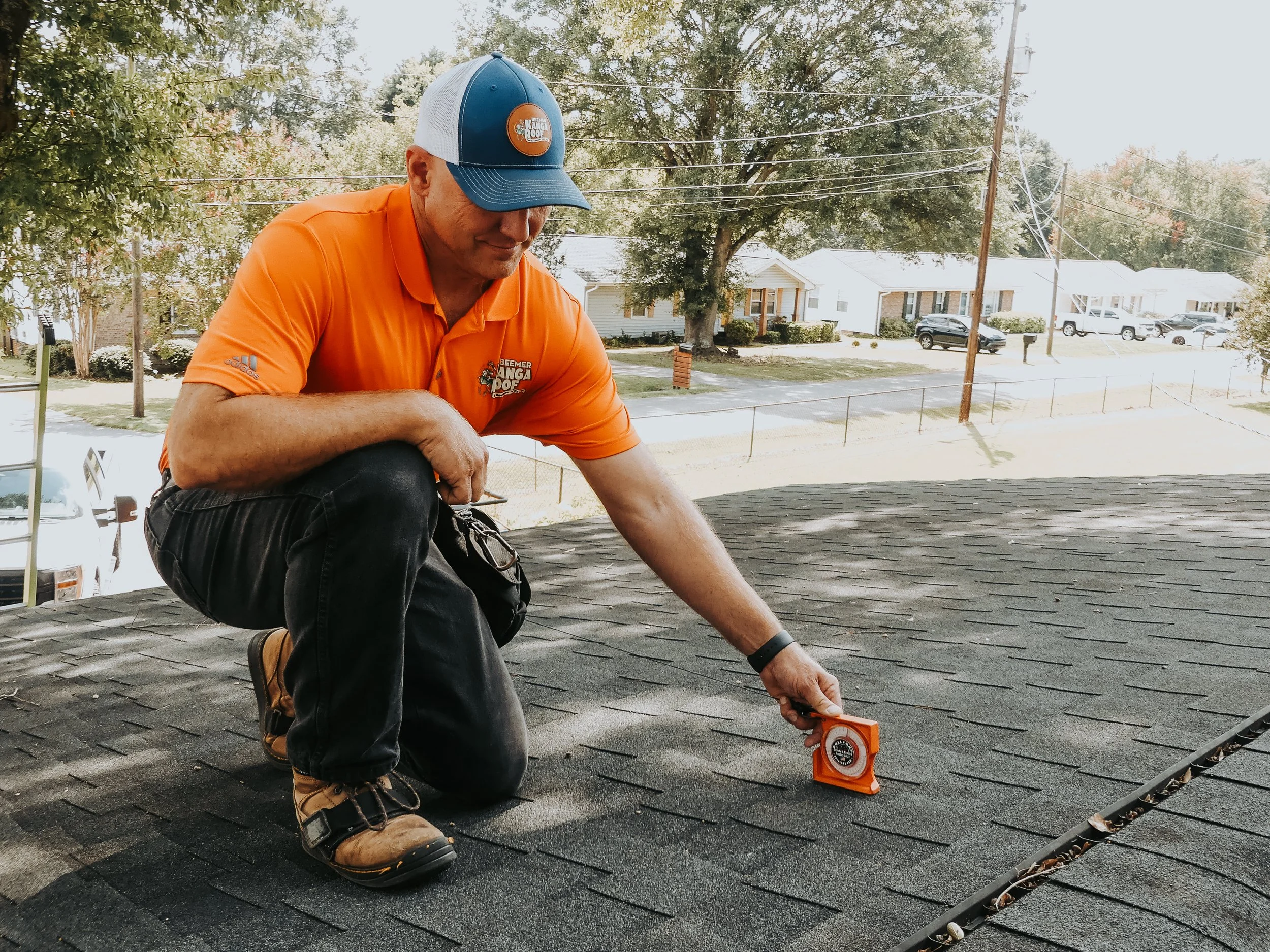 A man kneeling on a roof, measuring roofing shingles with a tape measure, wearing an orange polo shirt and a blue and white cap.