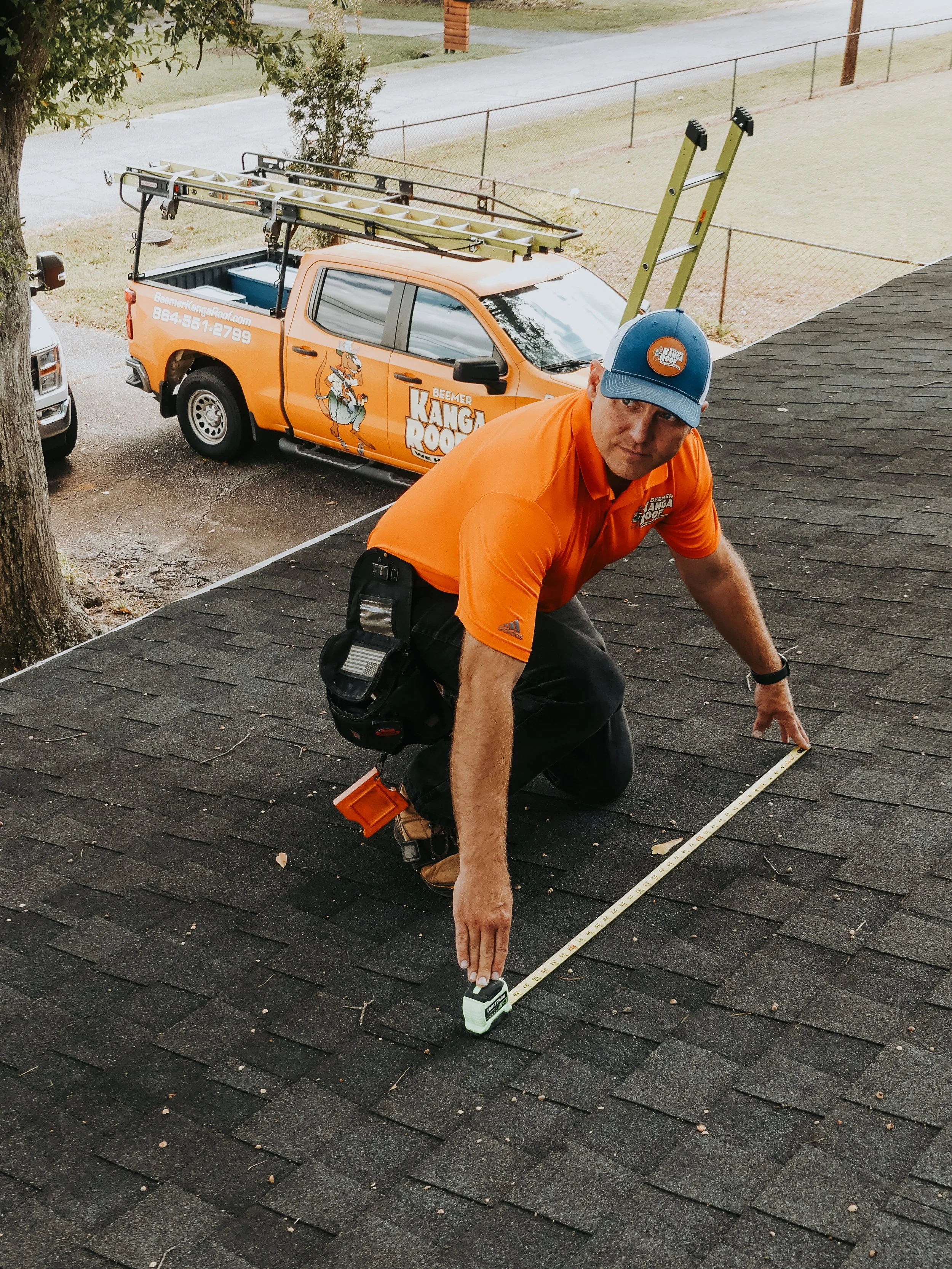 A worker measuring the roof of a house with a tape measure, wearing an orange shirt and blue cap, with a ladder and a work truck labeled 'Kanga Roof' parked nearby.