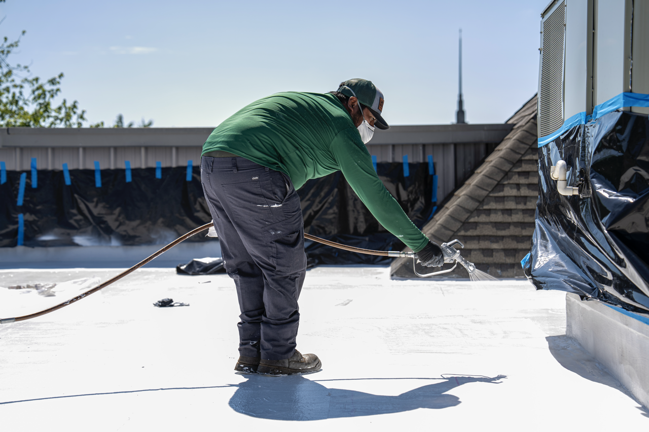 A man wearing a green shirt, black pants, a cap, sunglasses, and a face mask is spray painting a flat roof white.