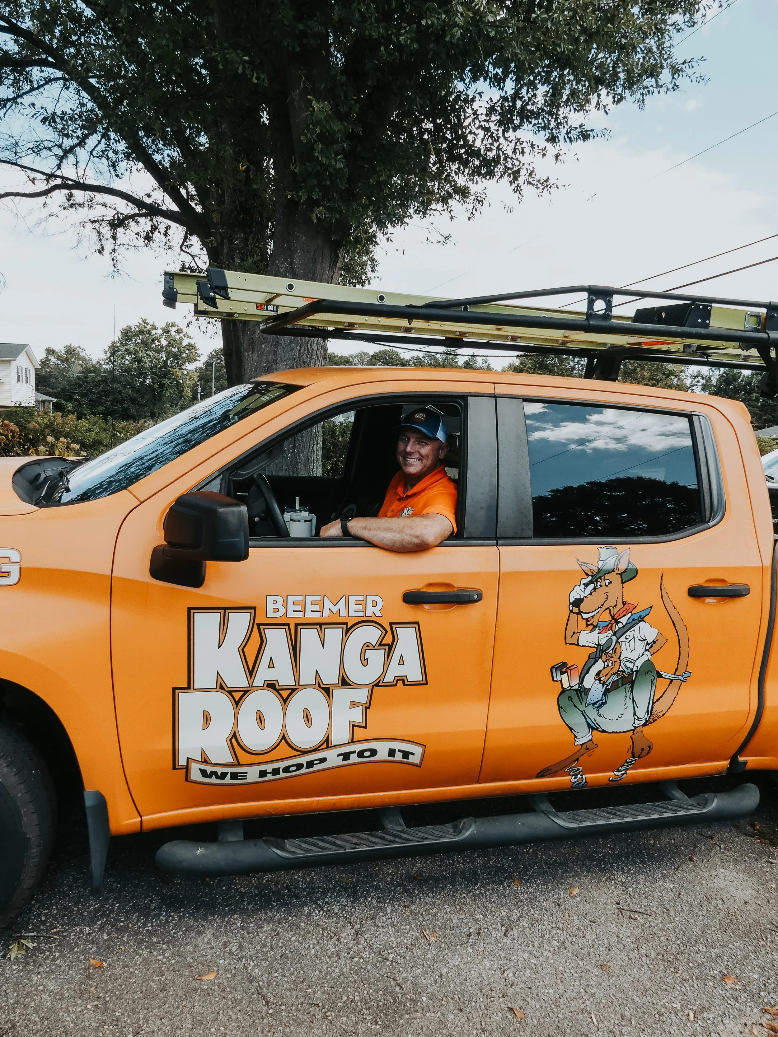 A smiling man in an orange shirt sitting in a bright orange truck with a ladder on top. The truck has a logo of an alligator dressed as a lumberjack with the words 'BEAMER KANGA ROOF' and 'WE HOP TO IT' on the door.