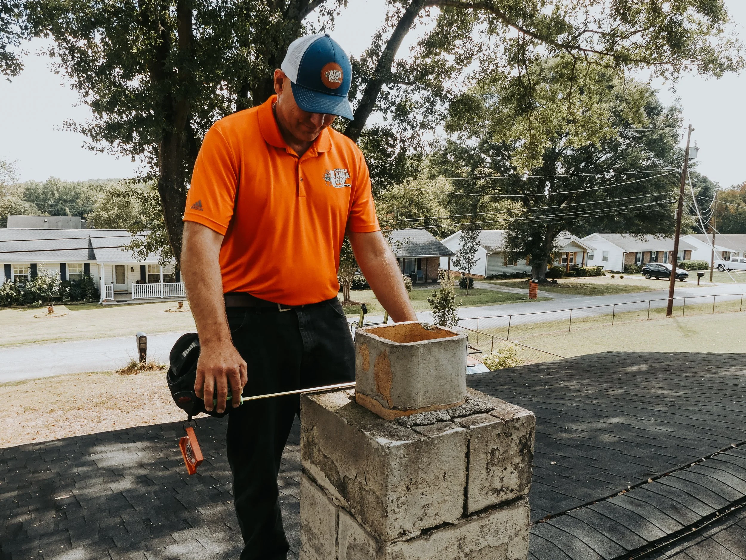 A man in an orange polo shirt and a baseball cap working on a chimney on a rooftop.
