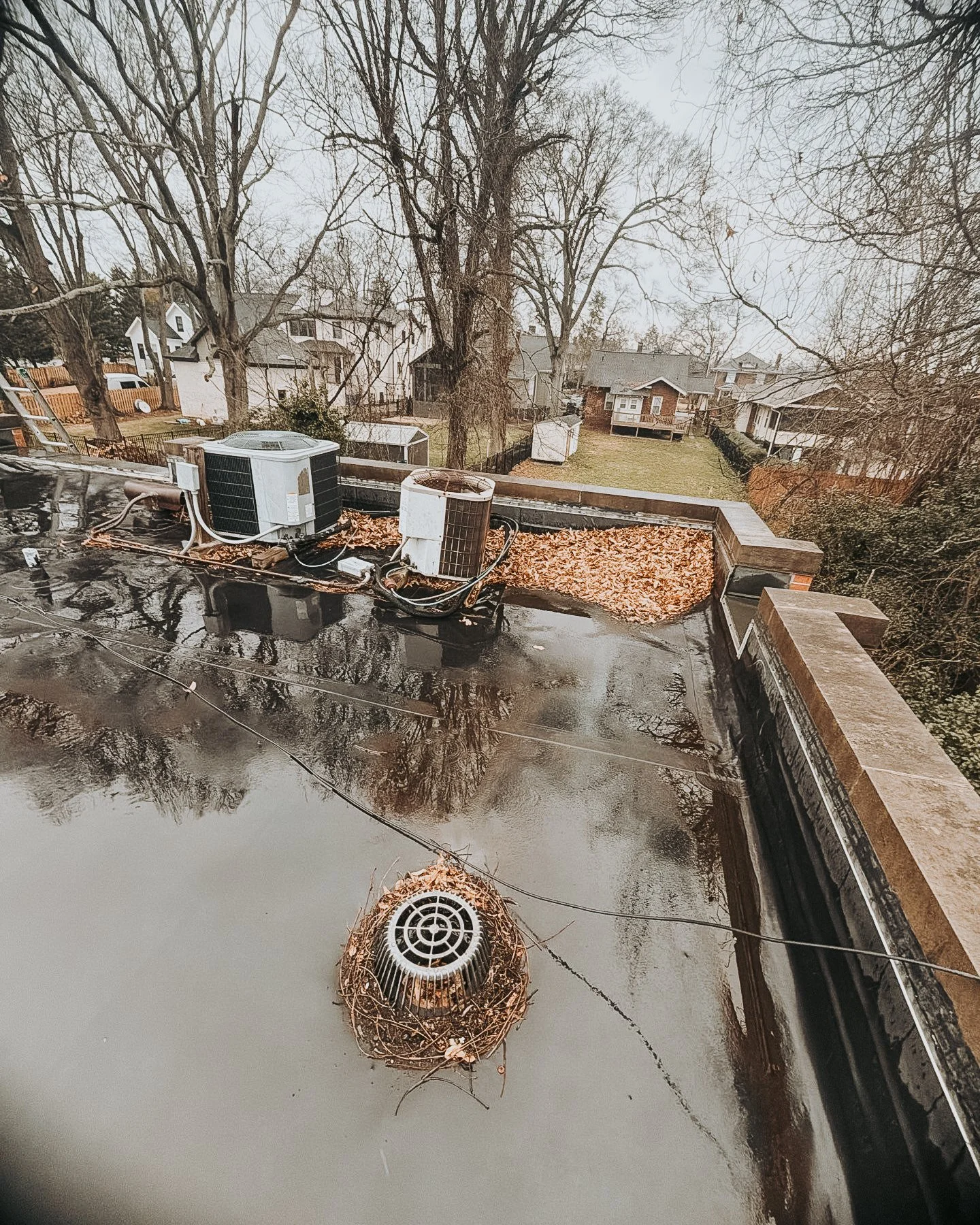 View of residential rooftops with an air conditioning unit and a vent, covered with leaves and water, overcast sky, trees without leaves in the background.