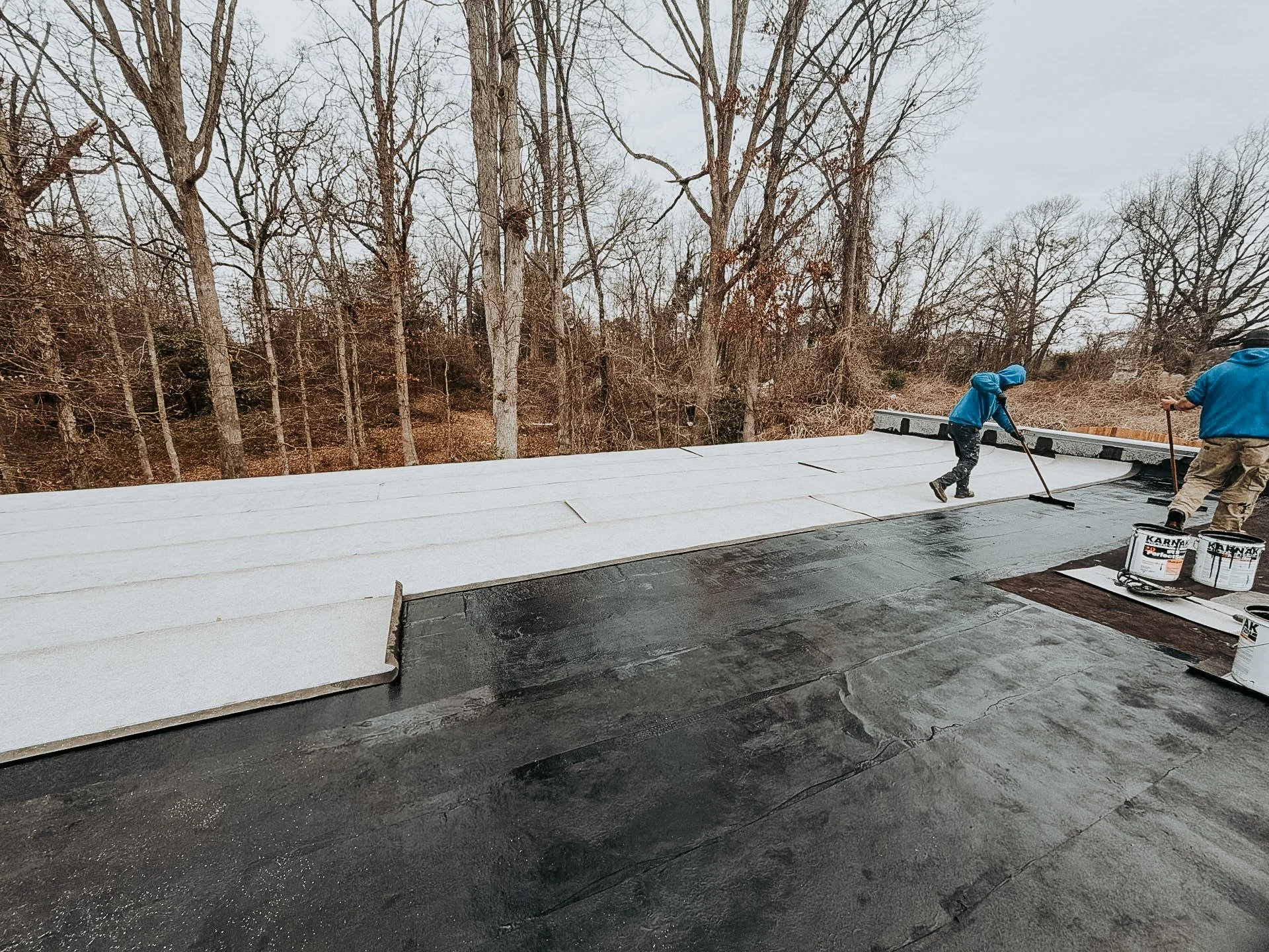 Two workers applying a reflective roof coating on a flat roof during daytime, with leafless trees in the background.