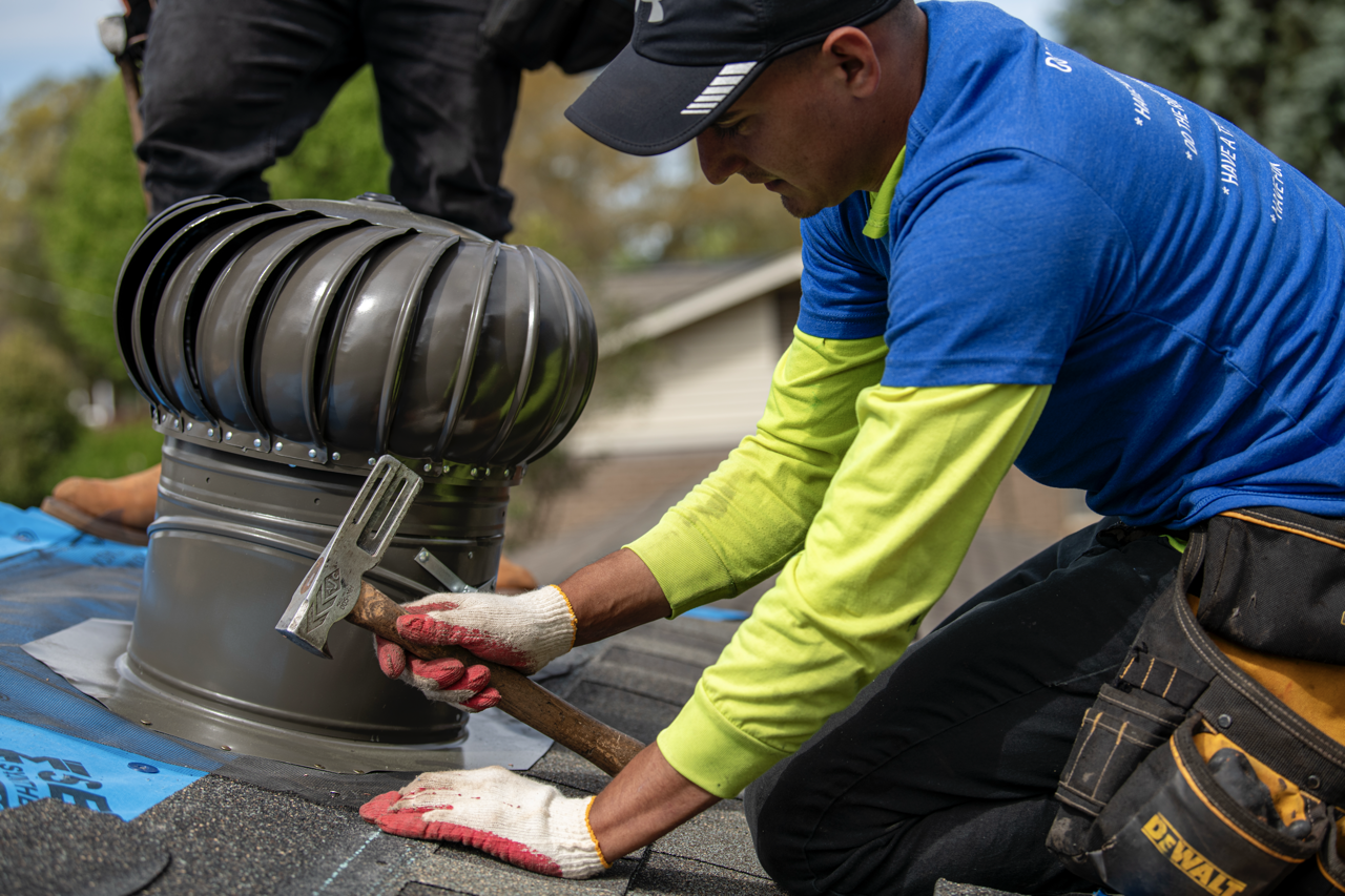 A man installing a roof vent on a house roof, wearing gloves and a blue shirt with tech gear around his waist.