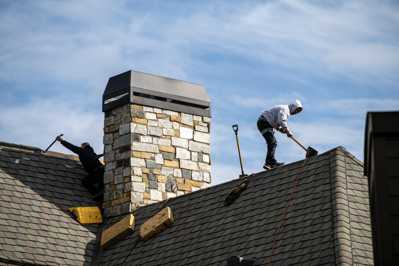 Two workers on a house roof replacing shingles, with a chimney in the center, against a blue sky with clouds.