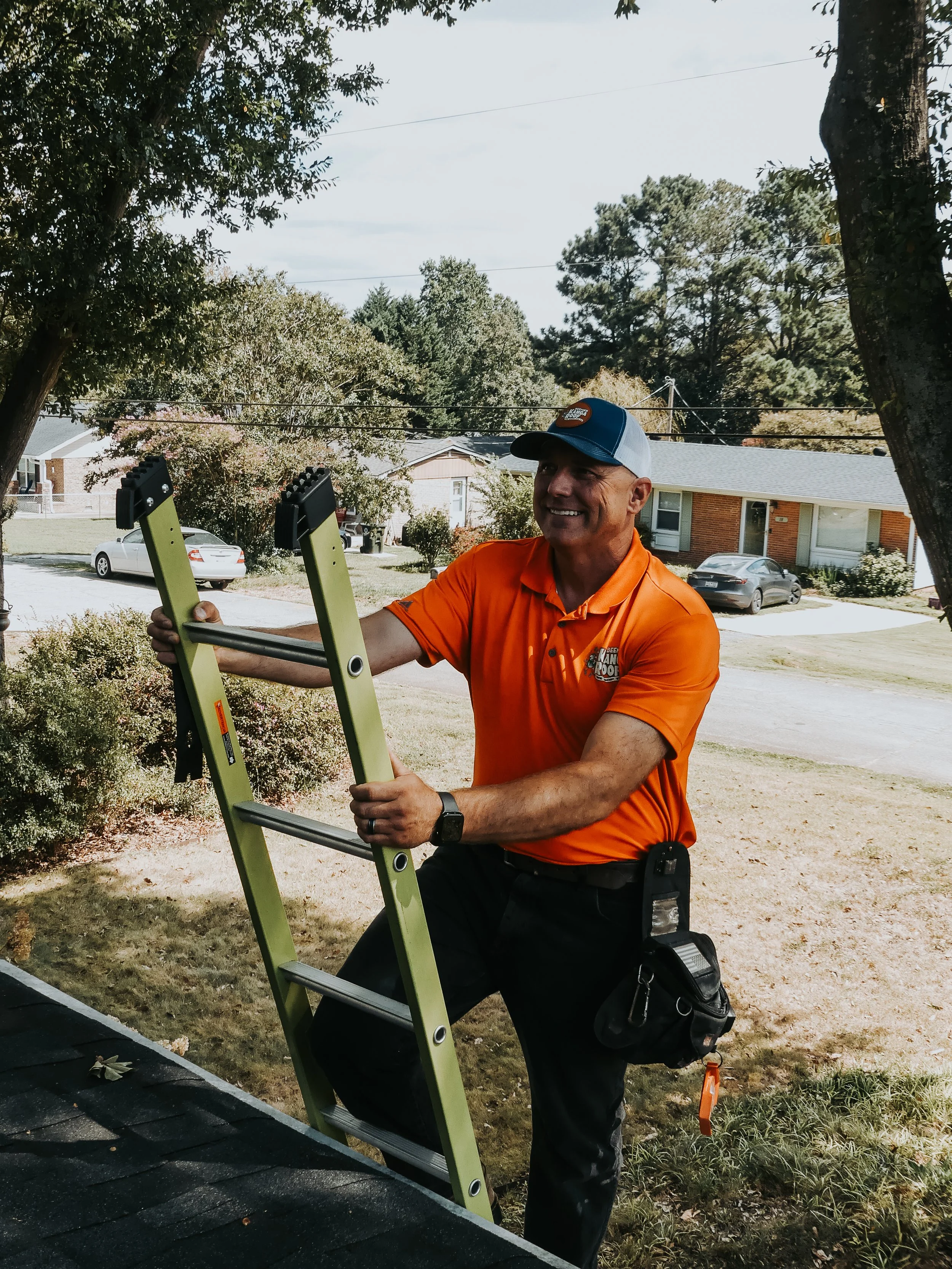 A man in an orange shirt and baseball cap standing outdoors on a ladder, smiling, near a residential neighborhood with trees and cars.