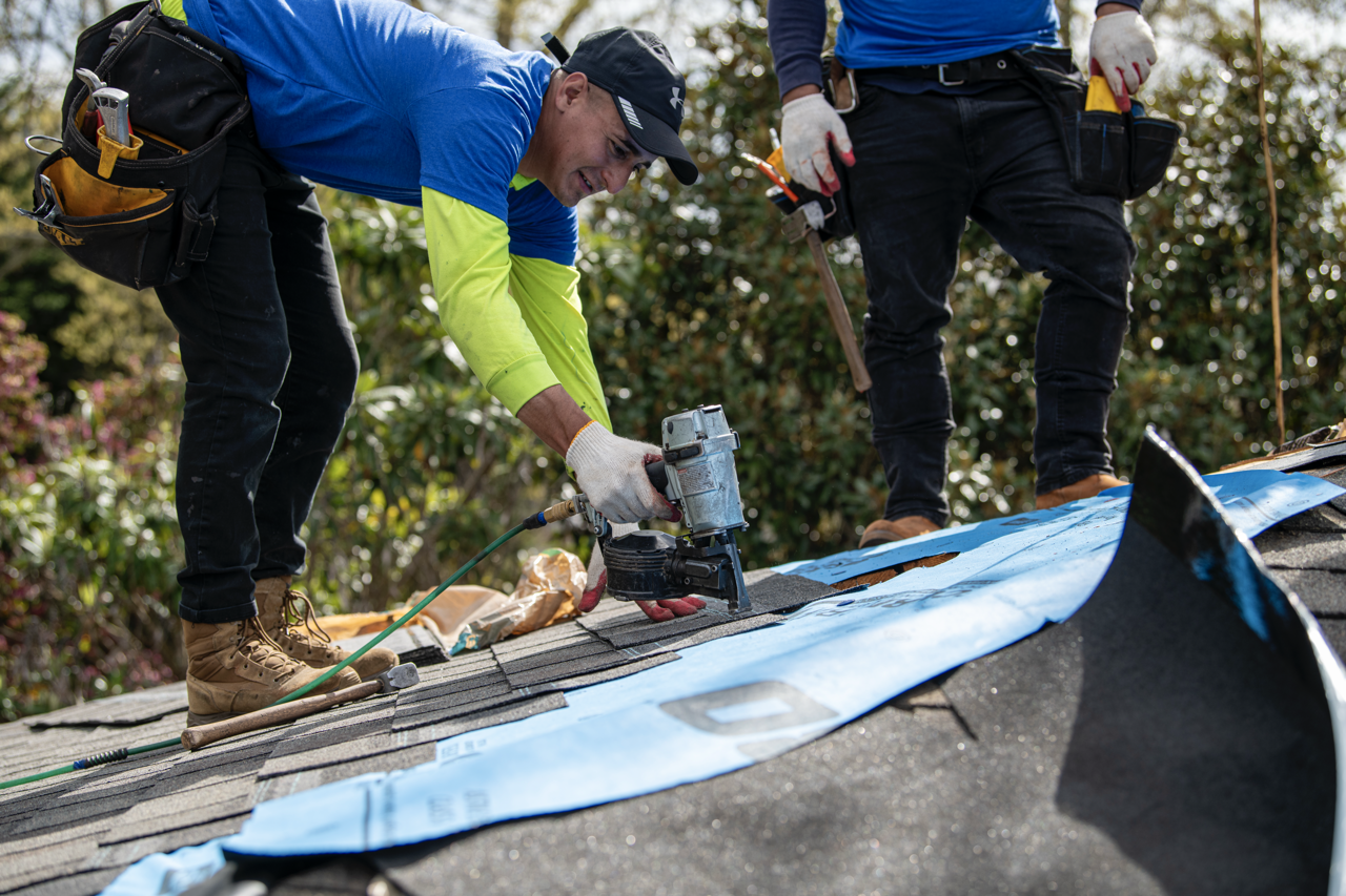 Two workers repairing a roof, one is using a nail gun while the other stands nearby with tools.