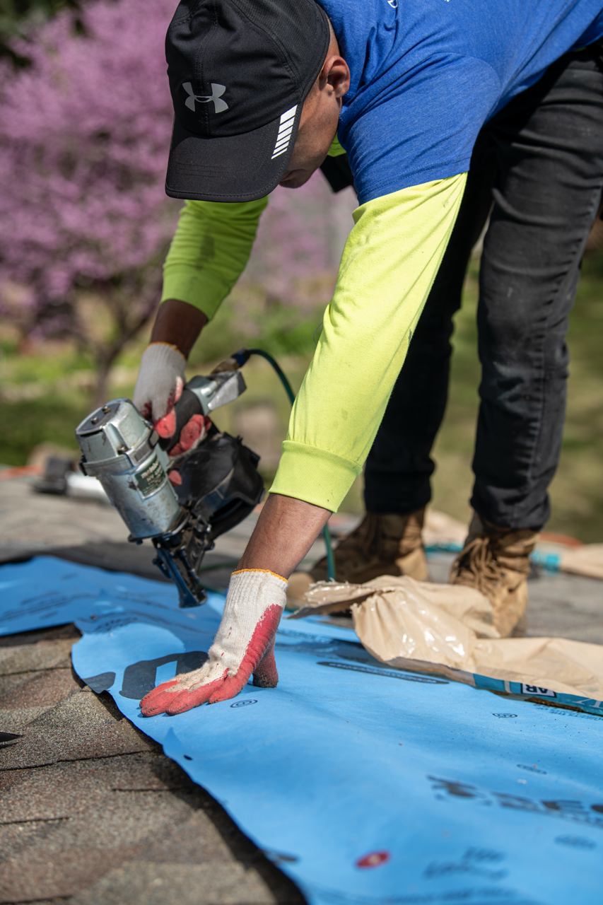 A person installing or repairing roofing on a house or building. The individual is wearing a black cap, yellow long-sleeve shirt, gloves, and is using a nail gun on blue roofing underlay, with pink flowering trees in the background.