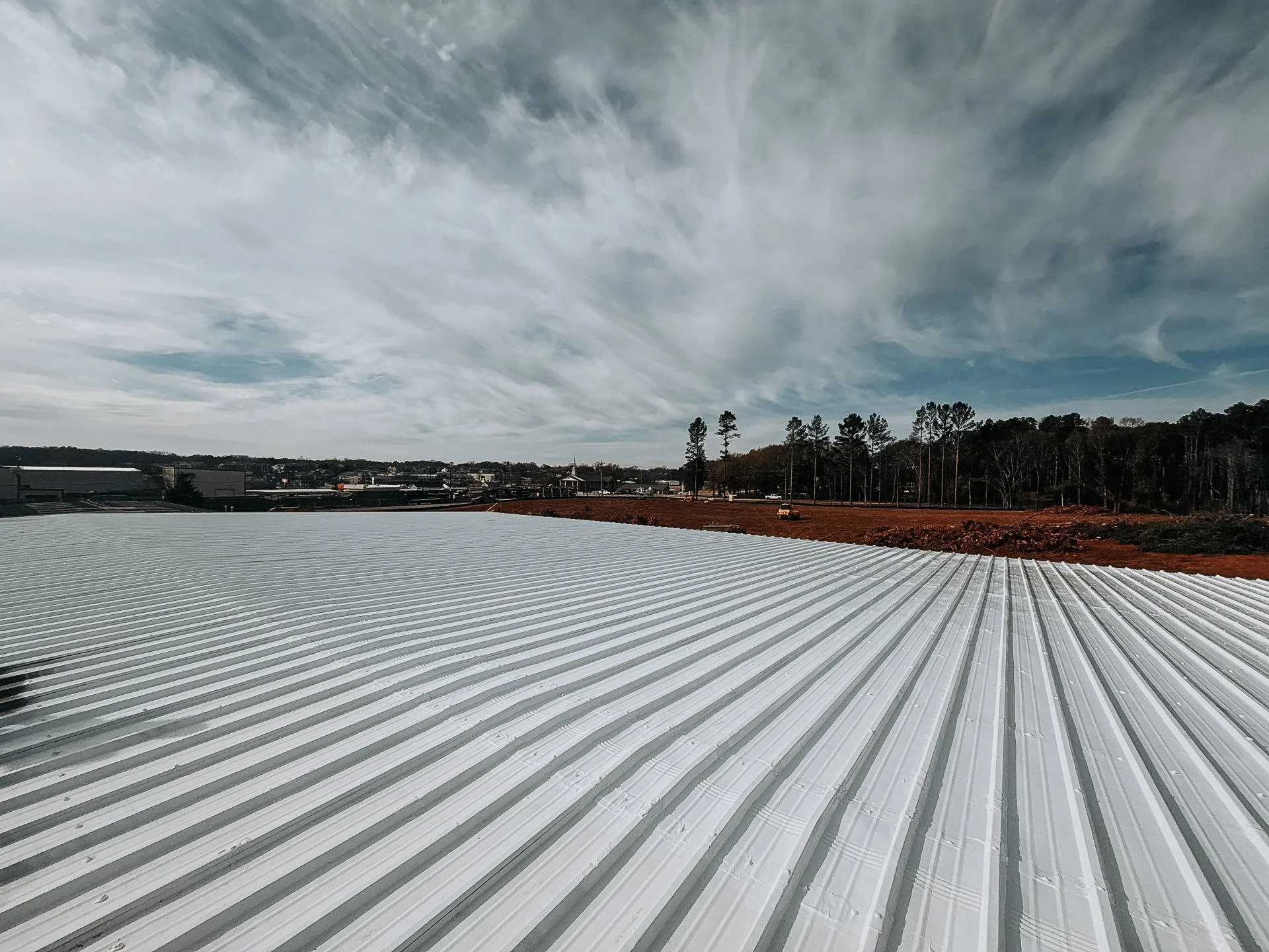 A wide view of a metallic corrugated roof with a rural landscape and trees in the background under a partly cloudy sky.