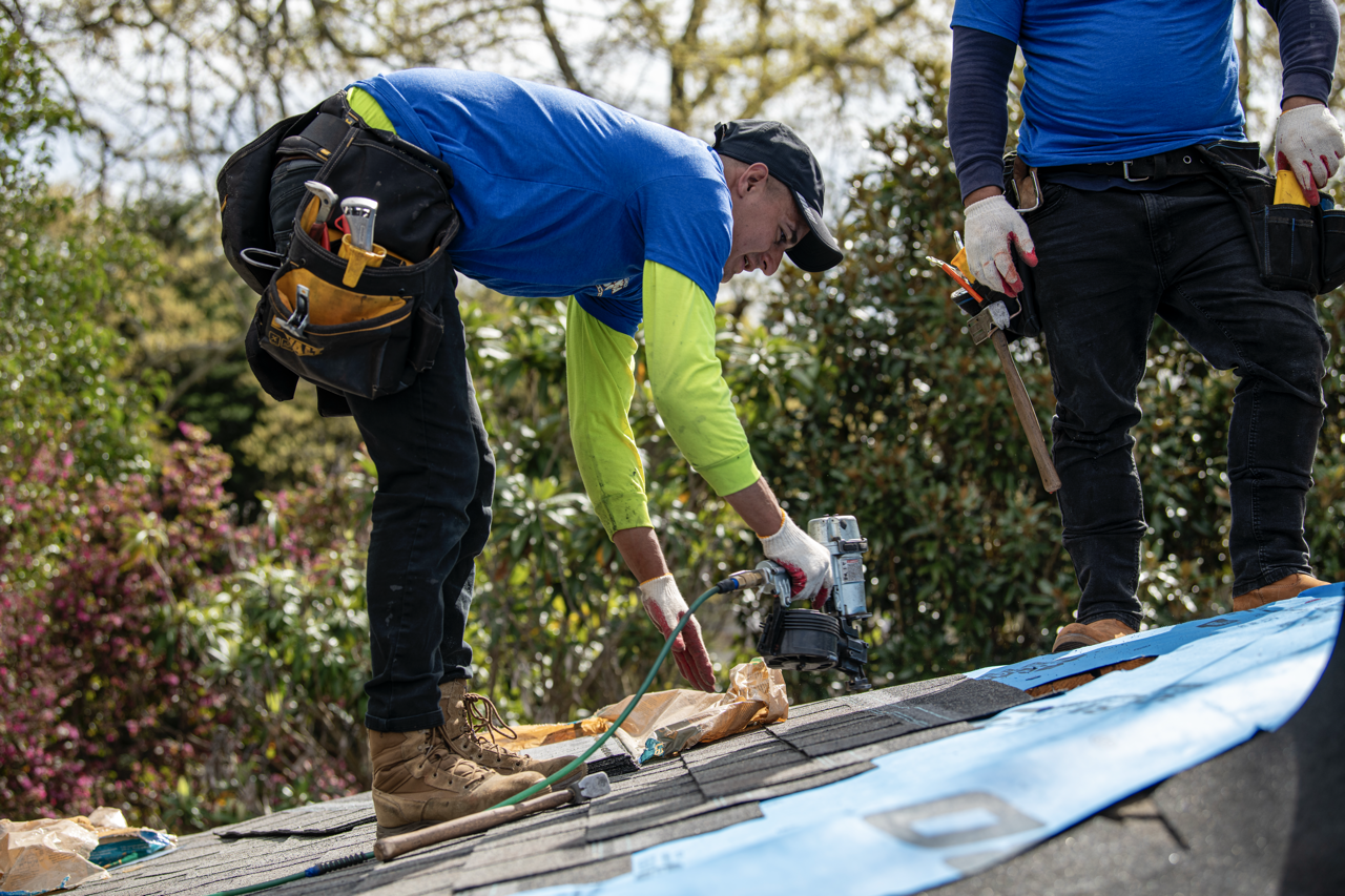 Two workers installing or repairing a roof outdoors, one is cutting or securing roofing materials while the other stands nearby.