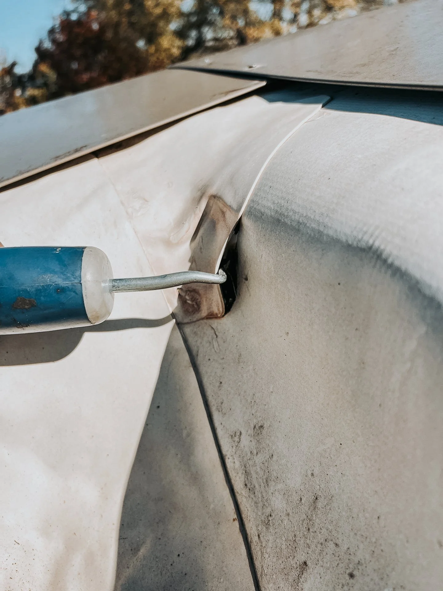 Close-up of a screwdriver being used to repair or modify a metal surface on a vehicle, with visible scratches and dirt.