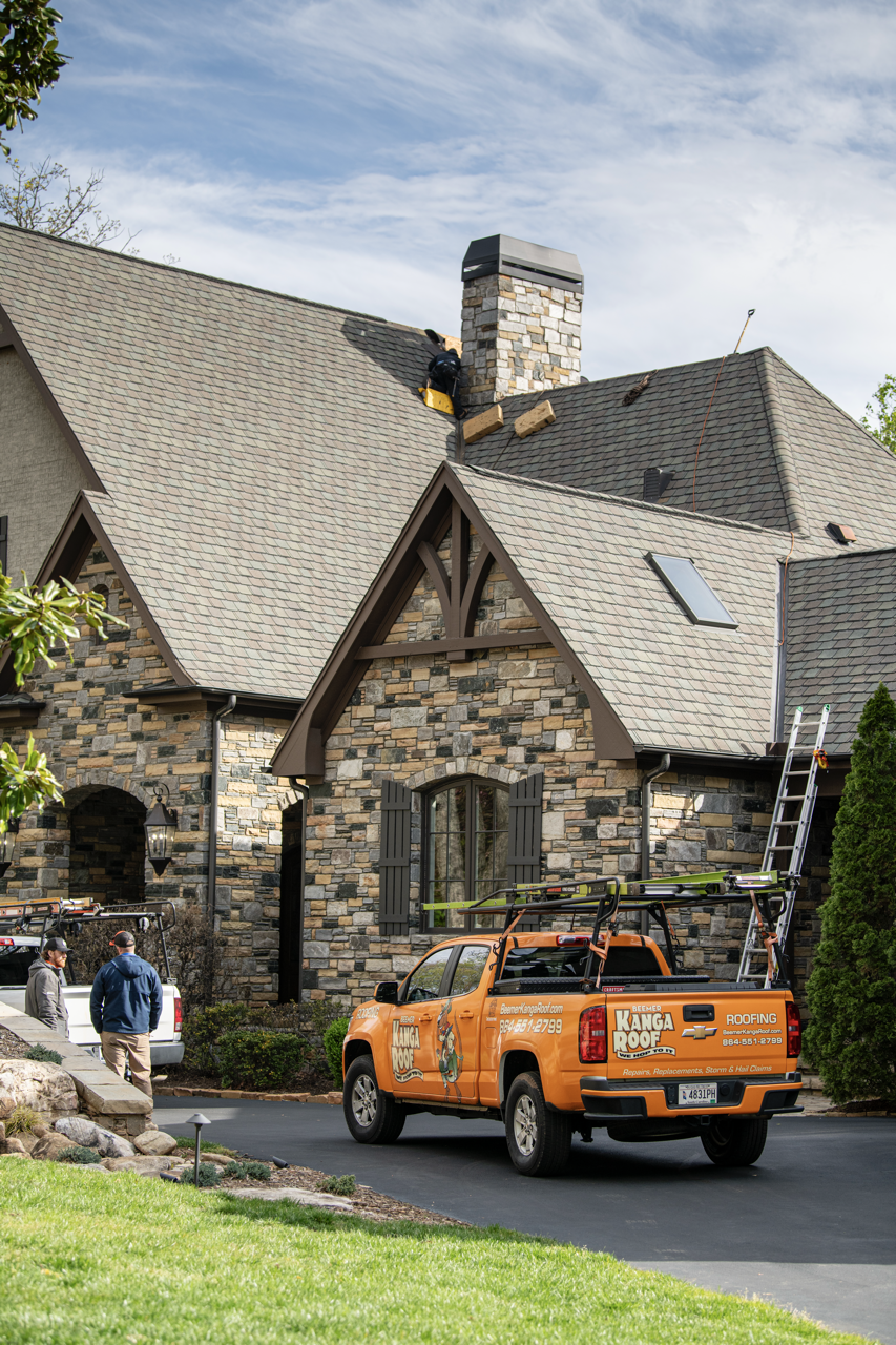 A roofing company worker installing a chimney cap on the roof of a large stone house, with two men standing nearby and a company truck parked in the driveway.