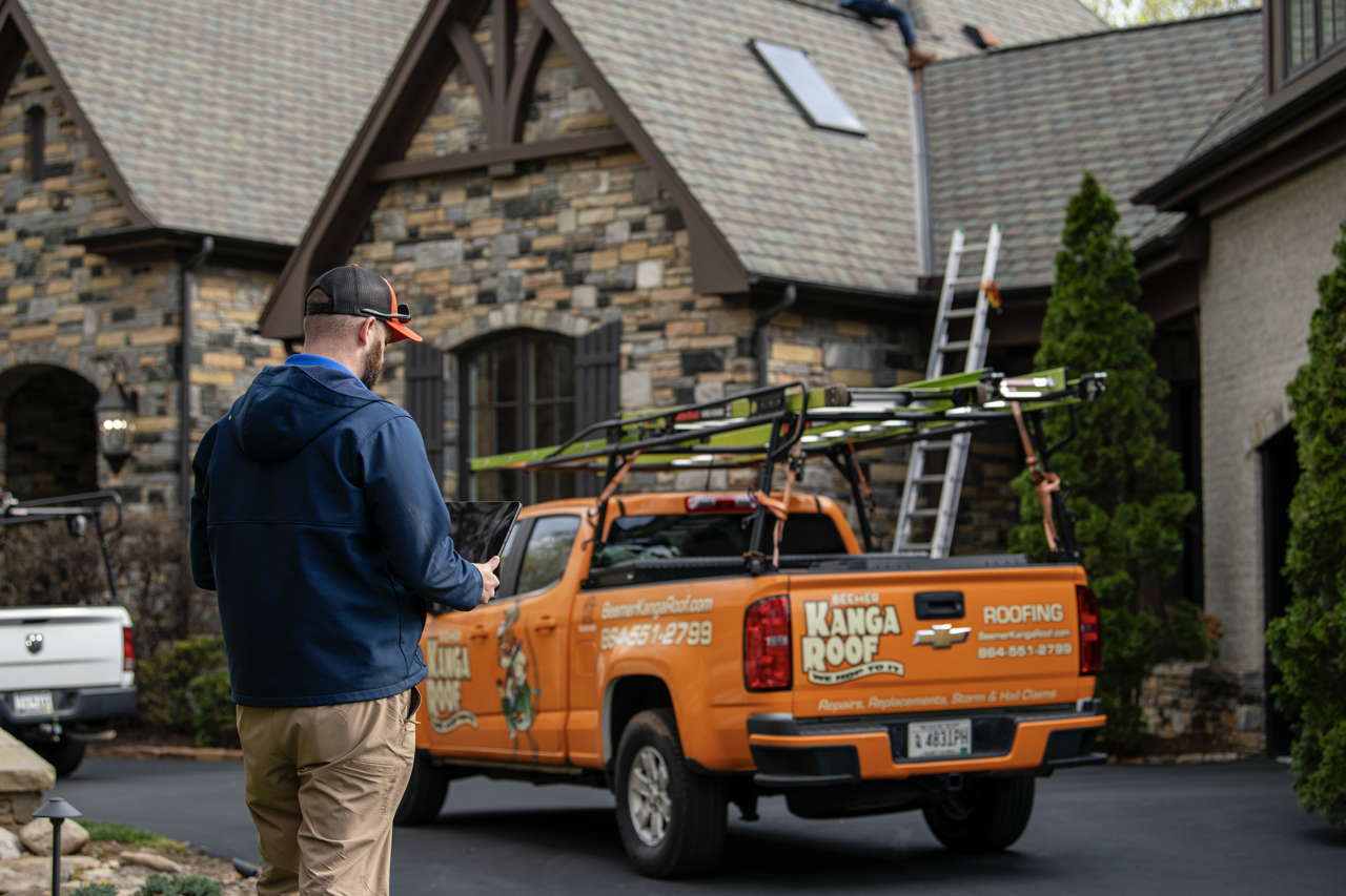 A man with a tablet standing in front of an orange roofing company's truck parked outside a stone house with a ladder leaning against the roof, indicating roofing work in progress.