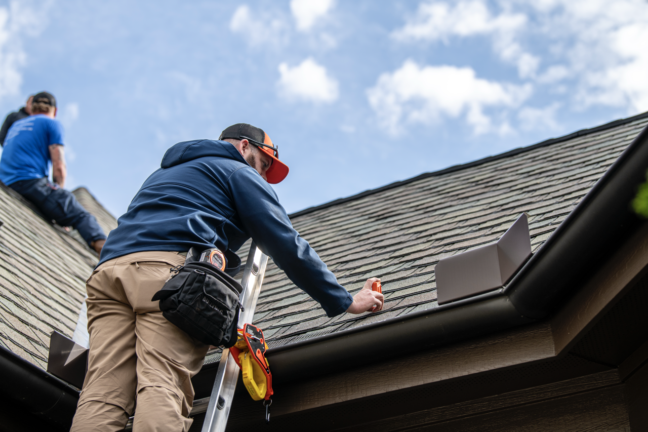 Two workers inspecting and repairing a roof on a house with blue sky and clouds in the background.