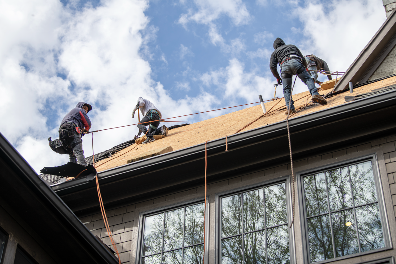 Three workers are reroofing a house, installing new wooden shingles on the roof while wearing safety harnesses and working at height.