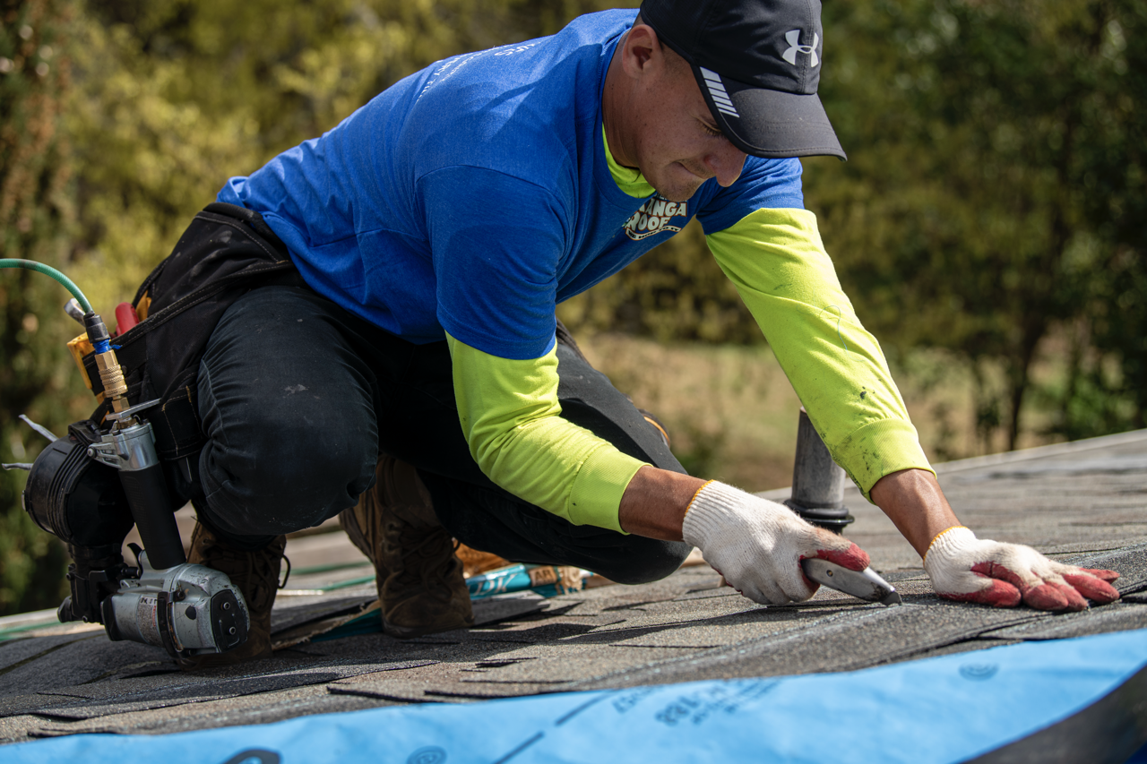 A worker wearing a blue and neon yellow shirt, black pants, gloves, and a cap is installing roofing shingles on a roof.