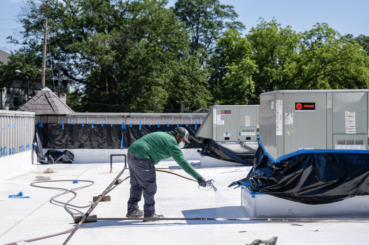 A worker in a green shirt and face mask applying a coating or paint on a flat rooftop with equipment and black tarps around.