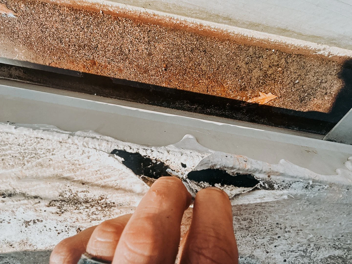 Close-up of a person's hand peeling worn paint from the edge of a window frame, revealing layers of old paint underneath.