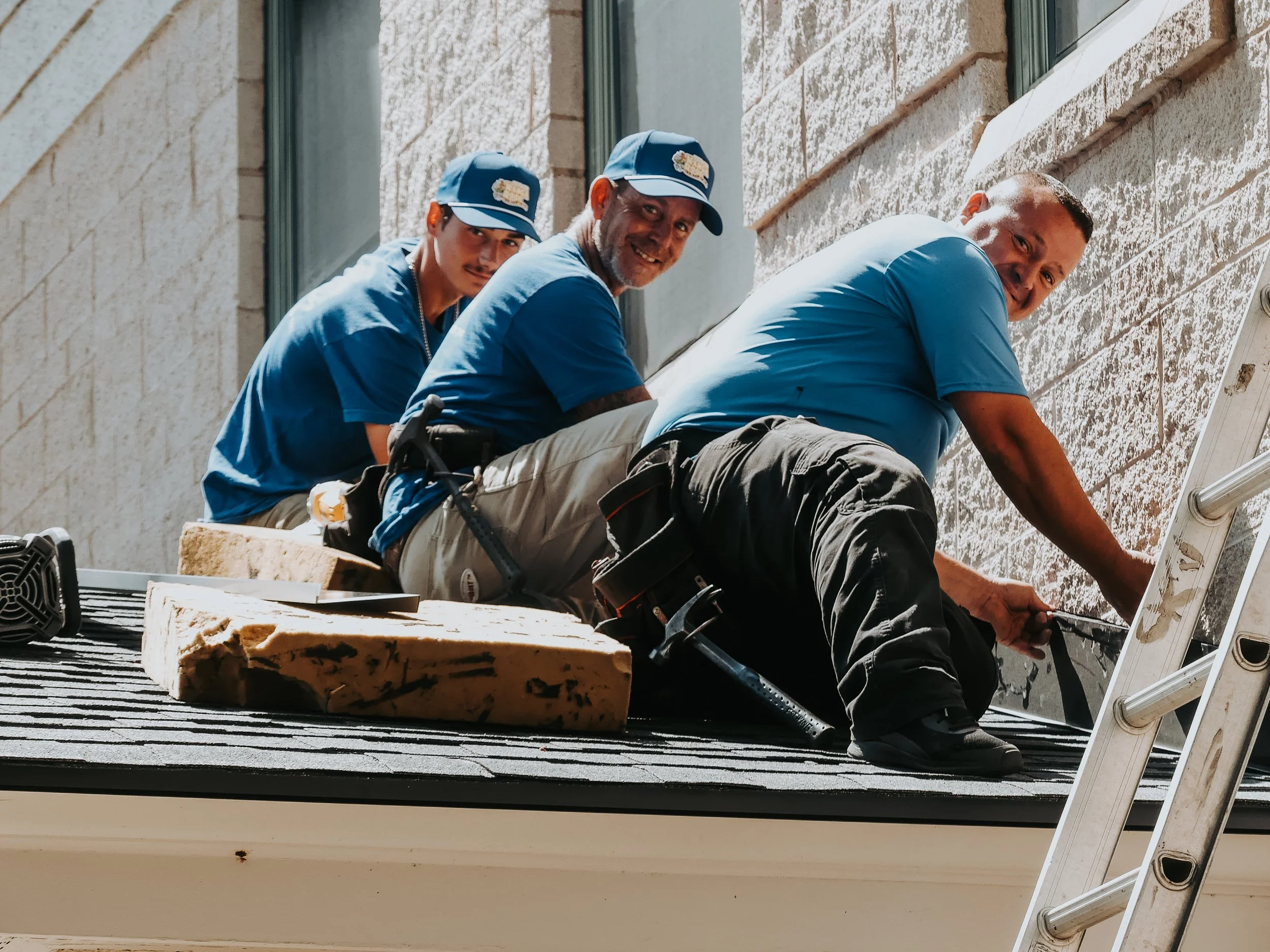 Three workers dressed in blue shirts and hats installing or repairing siding on a house exterior. They are on a roof with tools and material around them.