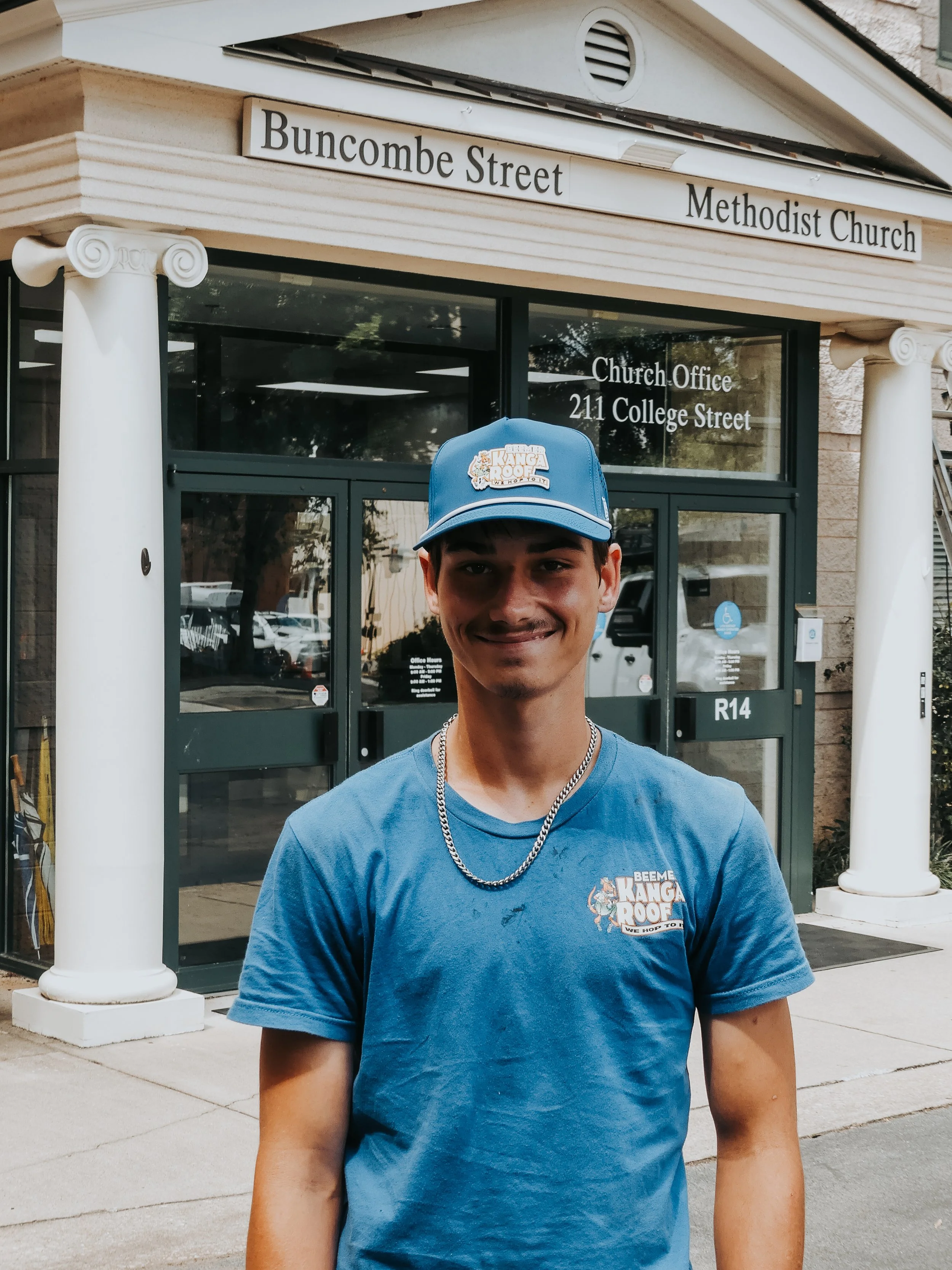 A young man wearing a blue shirt, matching blue cap, and a silver chain necklace, standing outside of the Methodist Church on College Street.