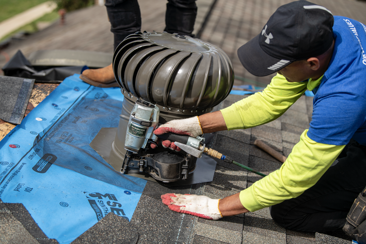 A worker installing a rooftop HVAC vent on a shingled roof, wearing a black cap, a blue and yellow long sleeve shirt, and gloves.