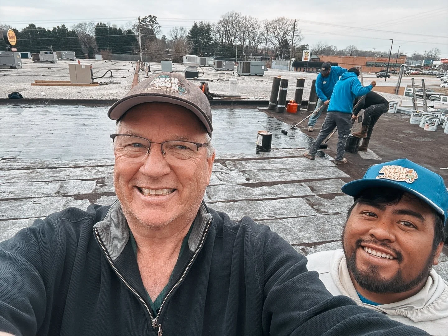 Two men taking a selfie on a rooftop during a roofing project, with workers in the background applying roofing materials.