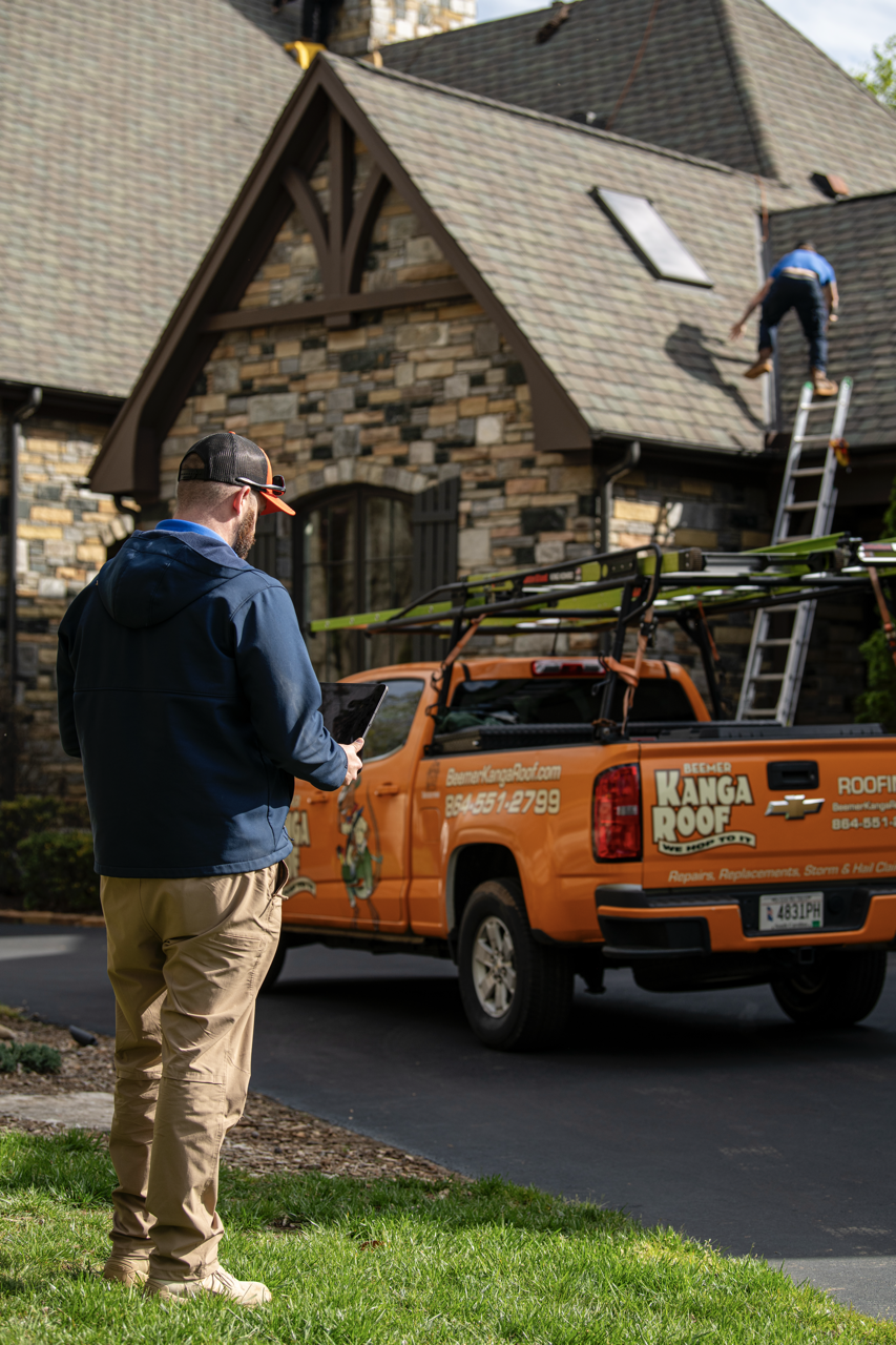 A roofer and a man with a tablet near a house with a steep, shingled roof. The roofer is working on the roof while the man observes. A bright orange roofing company truck is parked nearby.