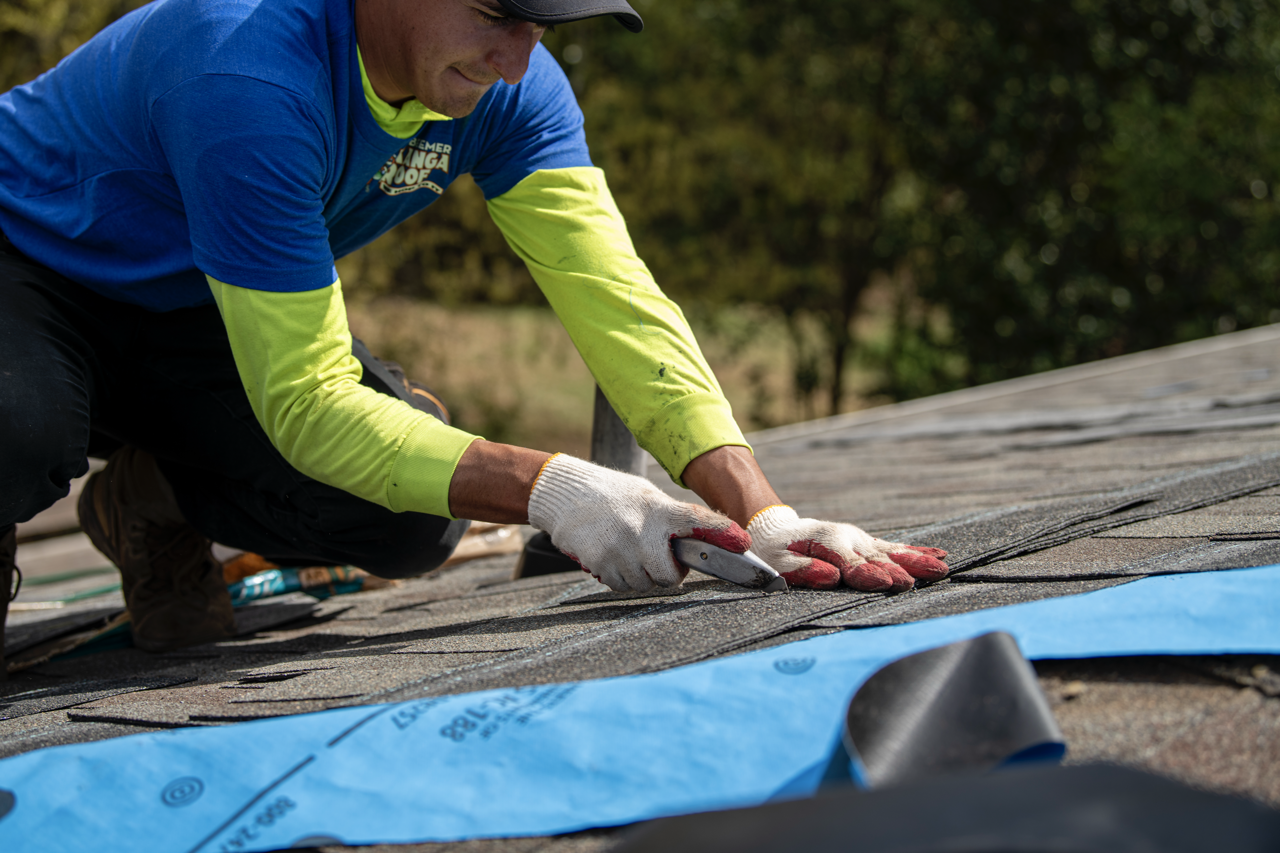 A person in work clothes installing roofing shingles on a roof, using a utility knife to cut the shingles.