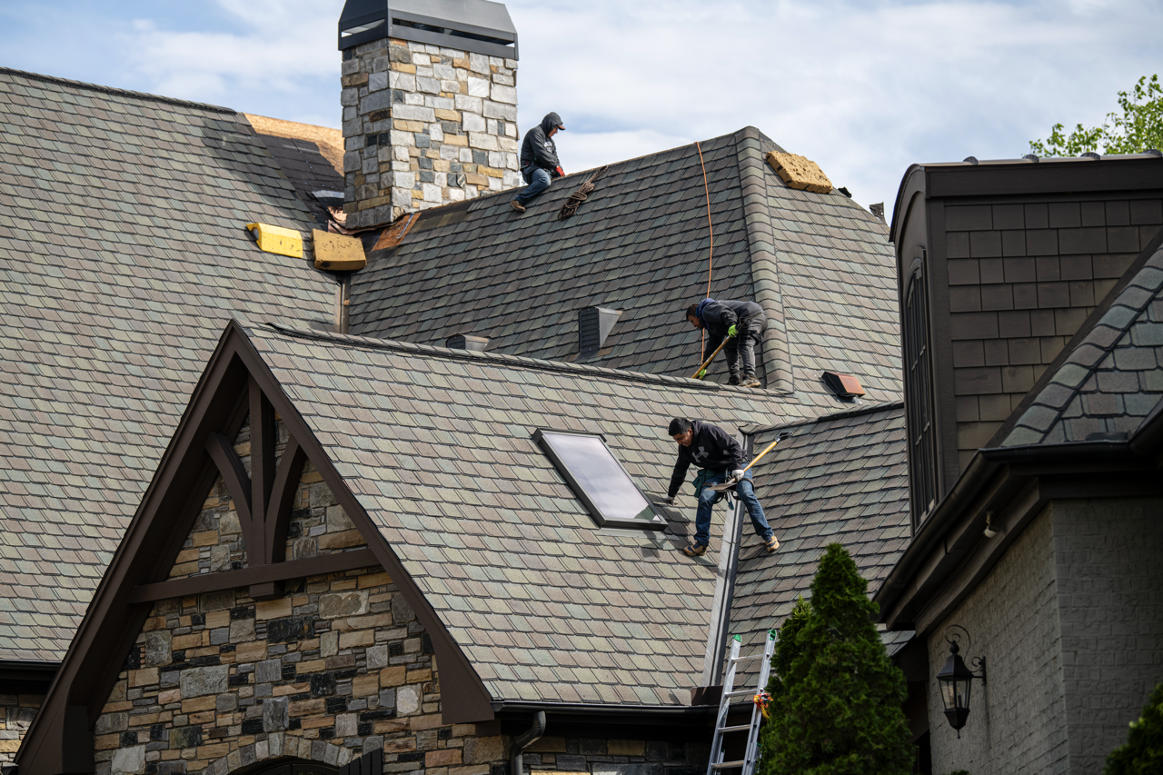 Three workers replacing a roof on a house with multi-colored shingles and a stone chimney. One worker is on the roof near the chimney, another is using a tool near the skylight, and the third is on the ladder working on the solar panel.