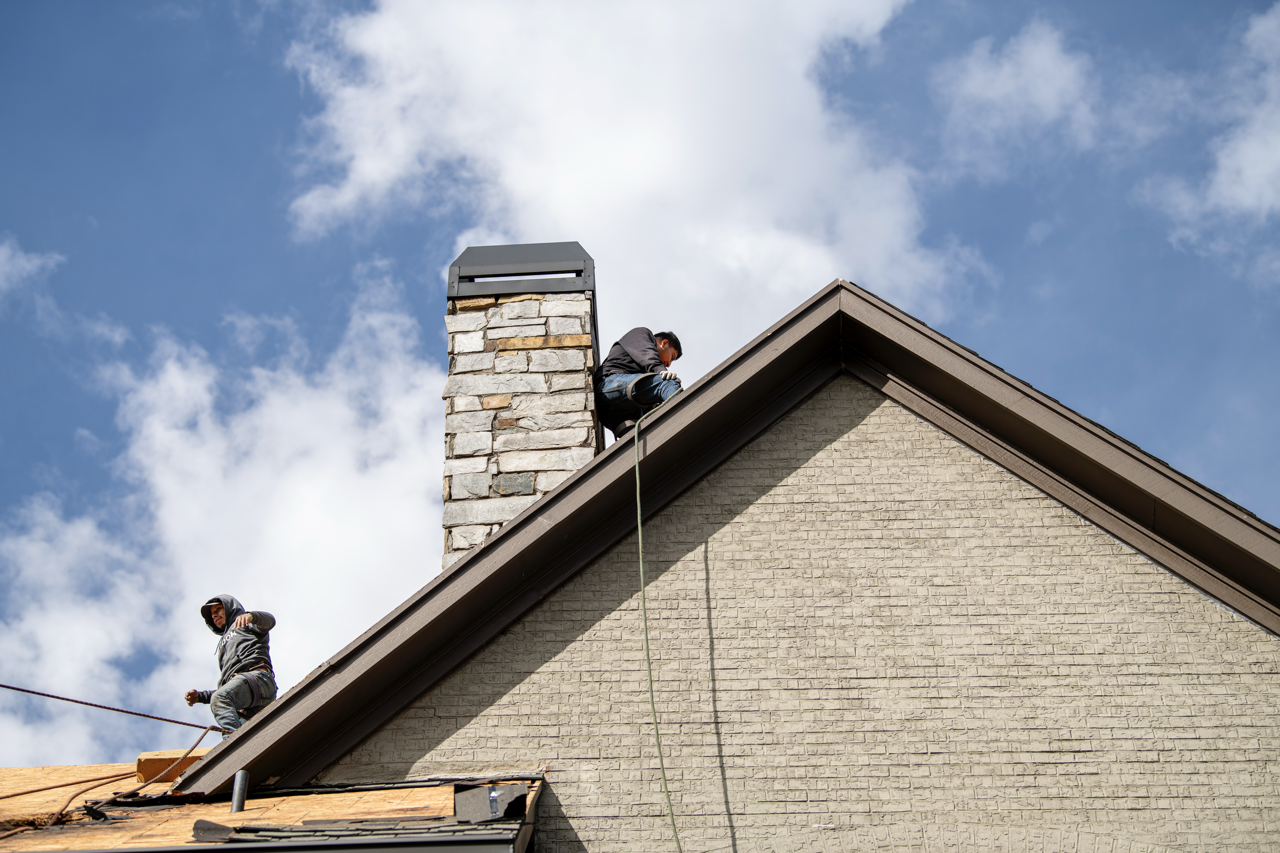 Two workers on a residential roof, one near the chimney and the other on the lower edge, installing or repairing roofing materials under a partly cloudy sky.