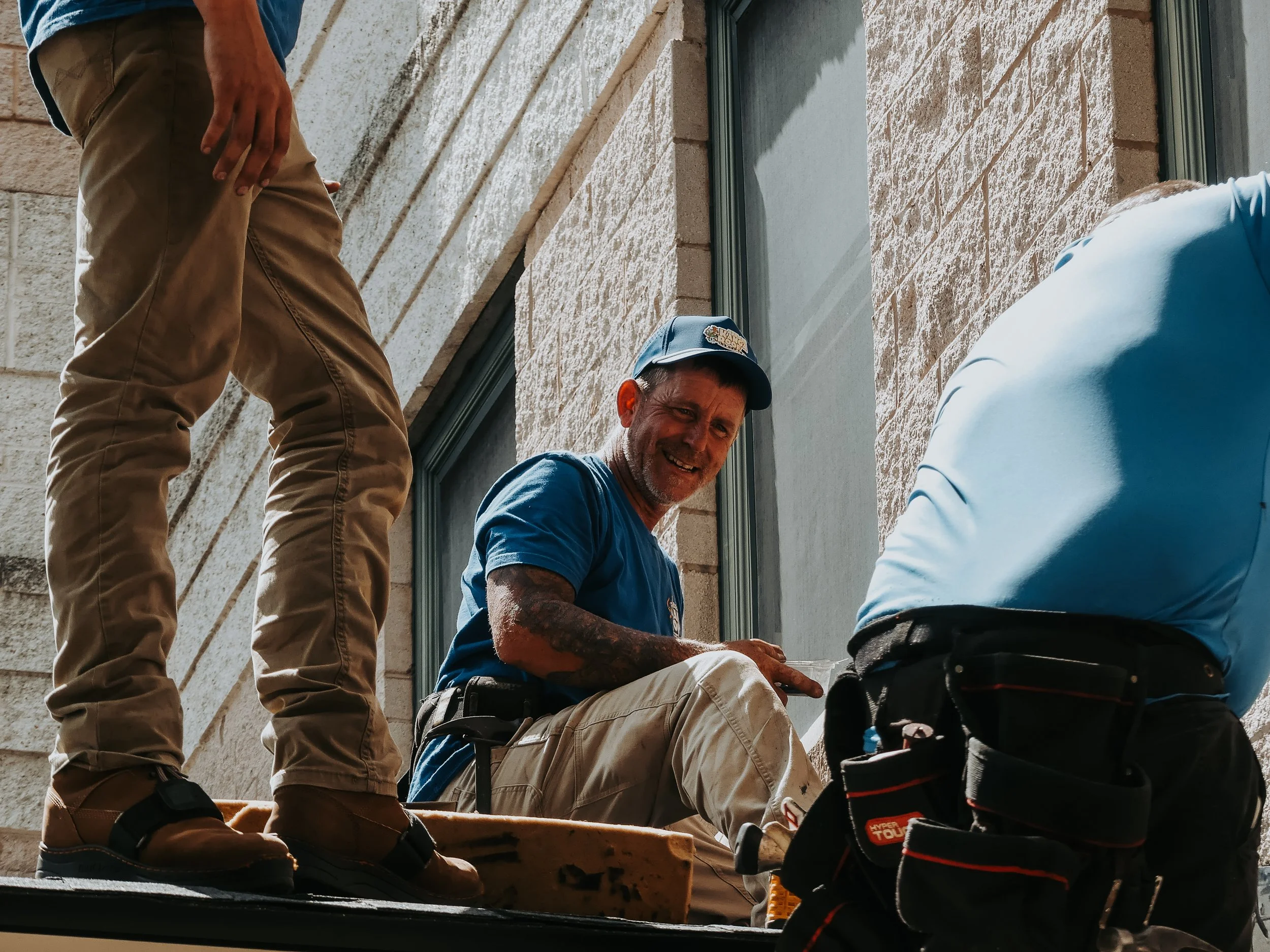 Construction workers installing a window on a brick building, with one man sitting and smiling, wearing a blue cap and shirt.